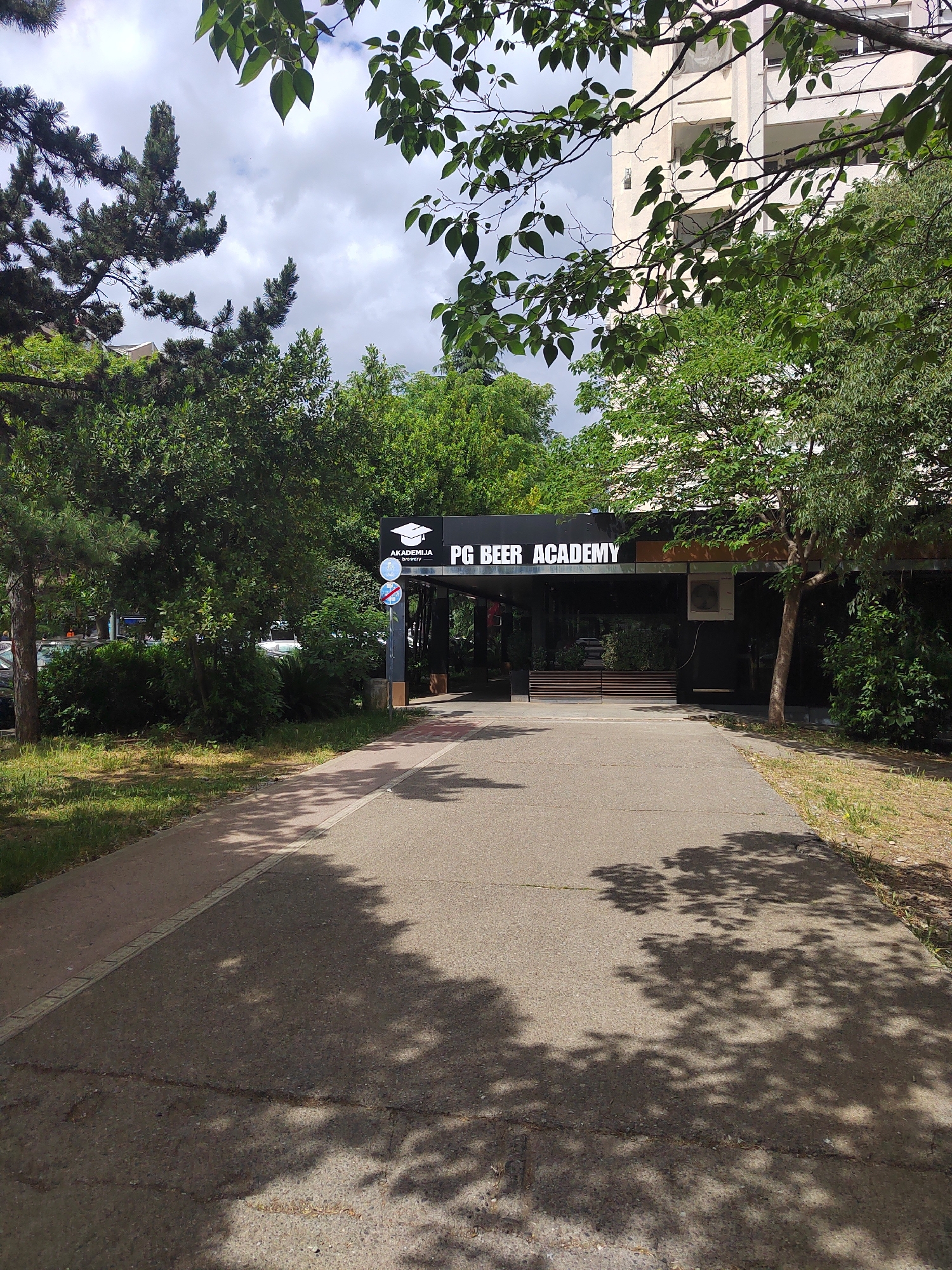 The image shows the exterior of "PG Beer Academy" in Podgorica, Montenegro. The academy has a black facade with glass windows and a sign with its name and a graduation cap logo. A paved walkway leads to the entrance, with trees and greenery surrounding the building. The sky is overcast. There are no people visible in the frame.
