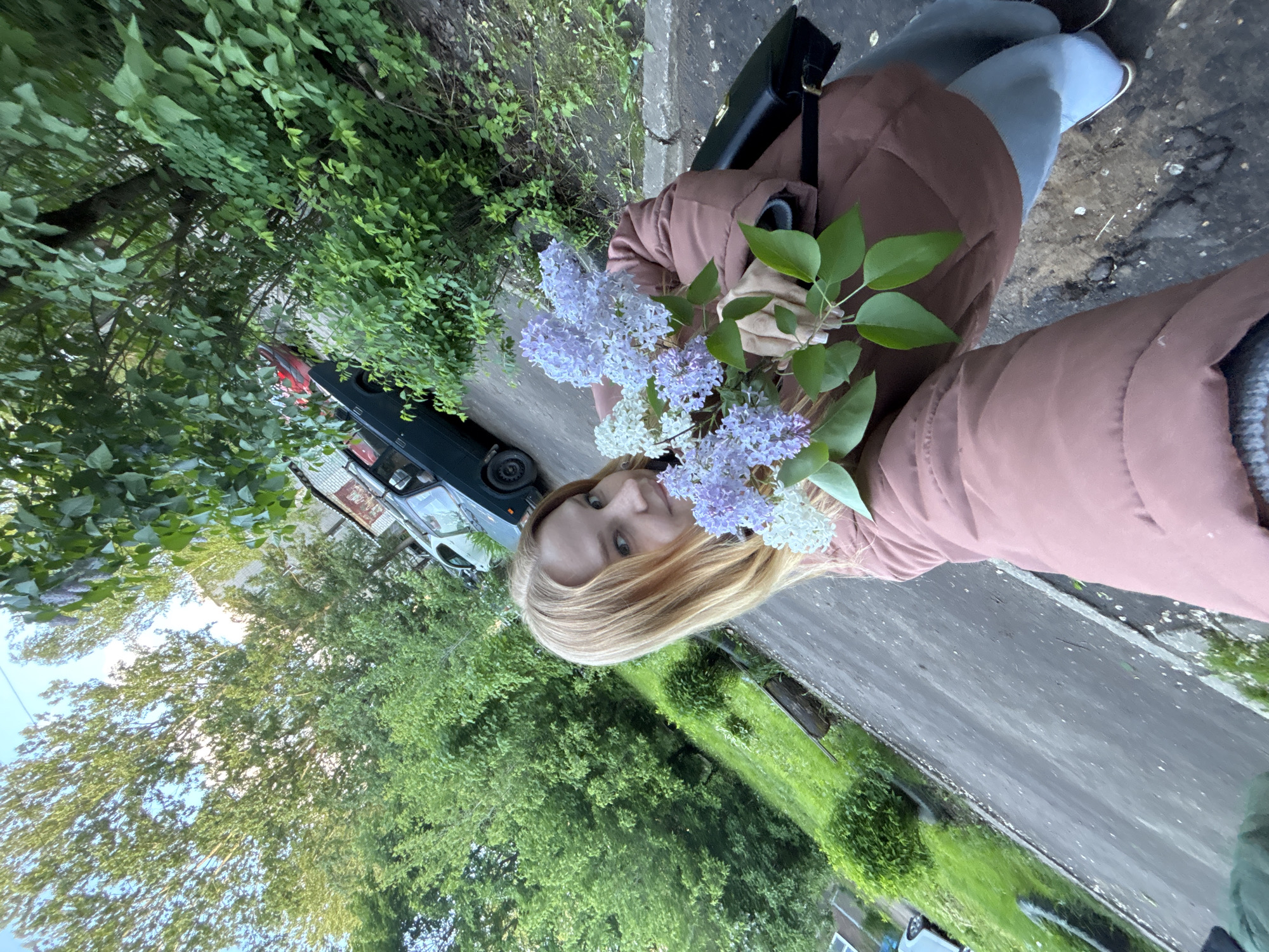 A young woman with blonde hair poses for a selfie while holding a bouquet of purple and white lilacs. She is wearing a pink jacket and jeans. Behind her, a dark green car is parked on the side of the road. The background shows a street in Rusanovo, Russia, lined with lush green trees and grass. A sidewalk runs along the road.