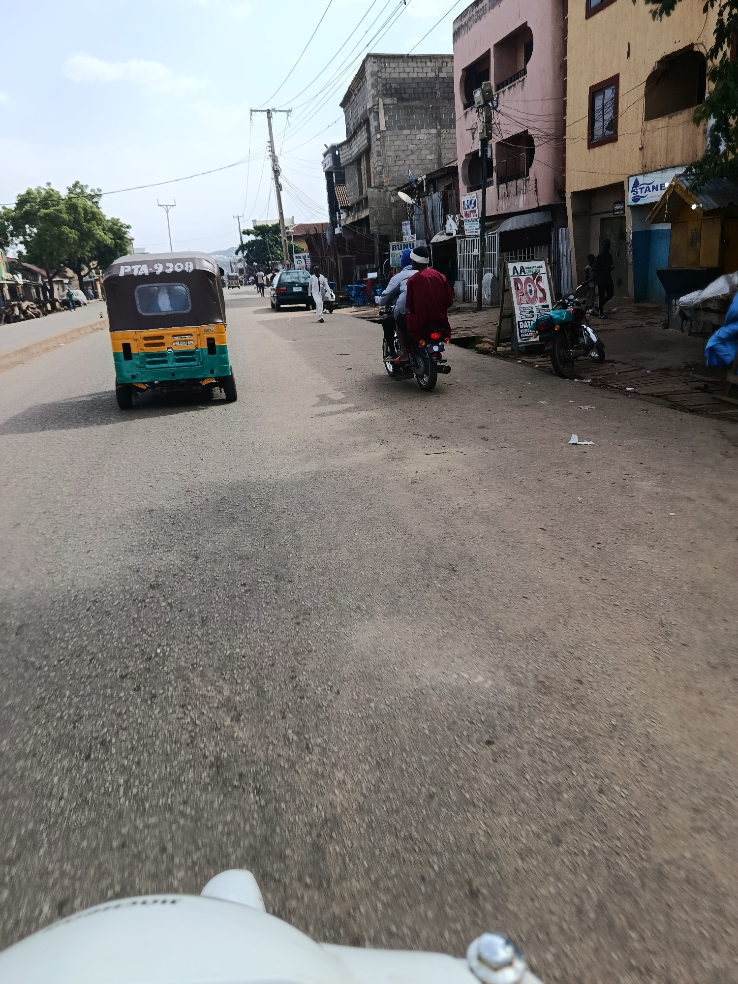 The image captures a street scene in Jos, Nigeria, likely during daylight hours under an overcast sky. In the foreground, the hood and headlight of a white vehicle are visible, suggesting the photo is taken from the perspective of a driver or passenger. The road surface is asphalt, showing signs of wear and tear.
Dominating the mid-ground is a three-wheeled auto-rickshaw (popularly known as a "Keke") painted in yellow and green, with the license plate "PTA-9308" clearly visible. The auto-rickshaw is stationary or moving slowly.
Further down the street, a motorcyclist wearing a red garment and a blue helmet is seen riding a motorcycle. Another person is on the back of the motorcycle. Several buildings line the right side of the street, varying in architectural style and color, some appearing older and some more modern. Signs indicating businesses, such as "STANES" and "AL-RAHEEM MODRAS," are visible on the buildings.
On the left side of the street, more buildings and what appear to be market stalls or roadside vendors are present, with trees providing some greenery. A dark-colored car is seen in the distance, also on the road. The overall impression is a busy urban environment with vehicular and pedestrian activity. The presence of overhead electricity wires and utility poles is characteristic of the infrastructure in many Nigerian cities.