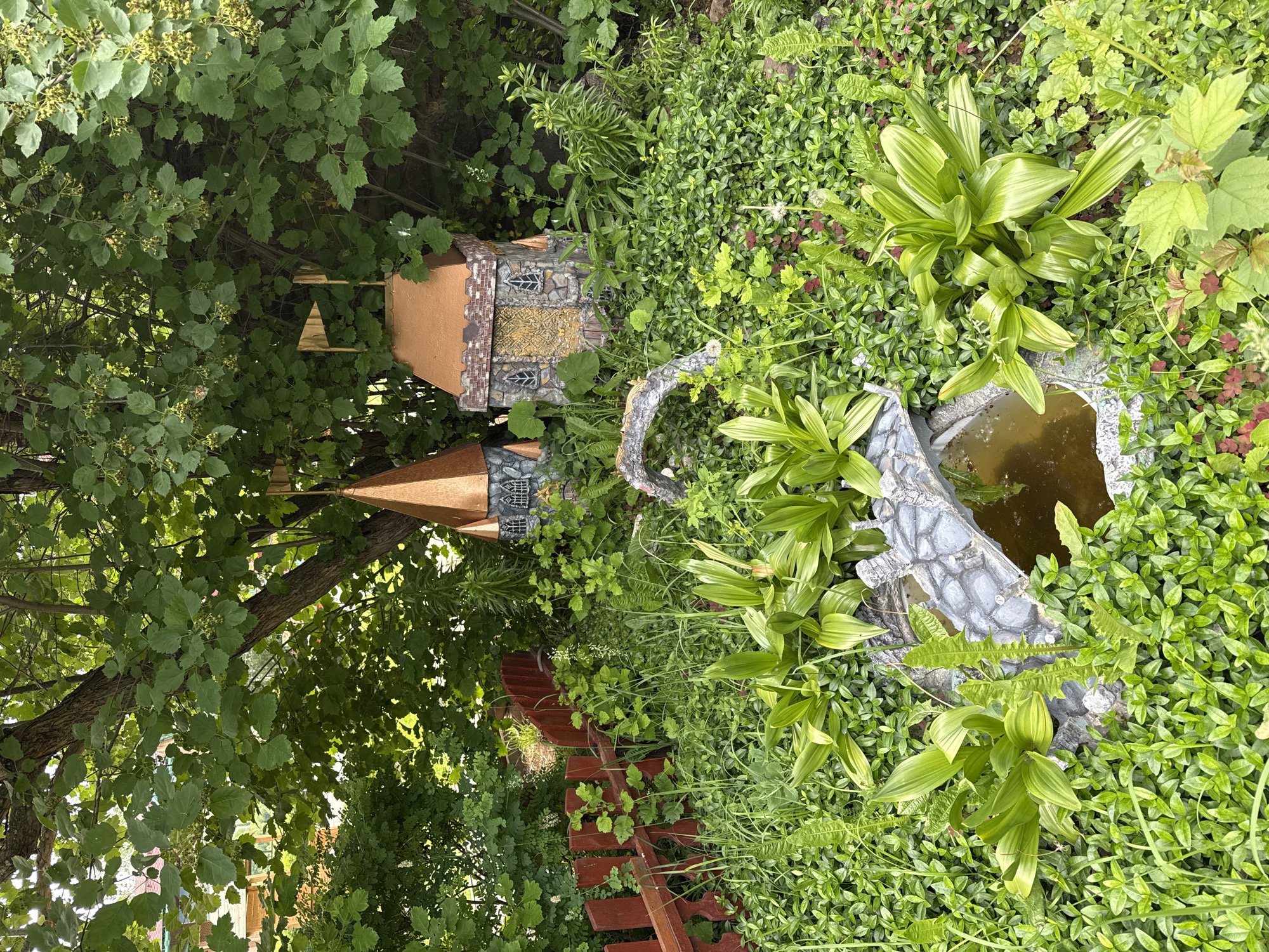 This image depicts a whimsical garden scene in Chaikovsky, Russia. In the center of the frame, two miniature fairy tale castles with copper roofs and stone-like facades are nestled amongst lush green foliage. A small arched bridge, also made to resemble stone, spans a shallow pool of water in the foreground. Surrounding the castles and bridge are various types of greenery, including ferns and ground cover plants. A red wooden picket fence runs along the left side of the image, partially obscured by the vegetation. The overall impression is one of a charming and slightly overgrown miniature fantasy world set within a natural garden environment.