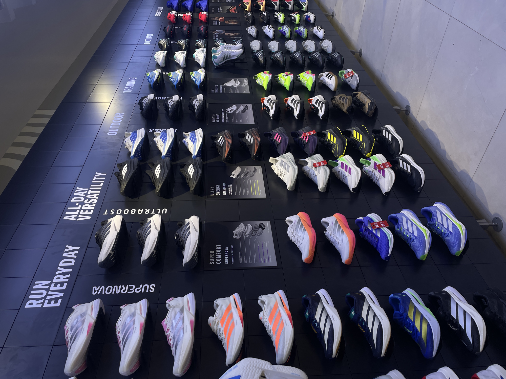 The image depicts an Adidas store interior, likely in Davao City, Philippines, showcasing a wide selection of athletic footwear displayed on a black wall. The shoes are organized by category, with headers such as "RUN EVERYDAY," "ALL-DAY VERSATILITY," "OUTDOOR," "TRAINING," "FOOTBALL," and "BASKETBALL." The shoes are predominantly Adidas models, with many featuring the brand's signature three stripes. Various colors and designs are visible, including running shoes with bright soles, training shoes, and football boots. Several shoes have price tags or promotional labels indicating discounts, such as "30% OFF." The lighting is bright, illuminating the products and the store environment. The floor is tiled in a neutral gray color. There are no people visible in the image, suggesting the store may be closed or the photograph was taken during a quiet period. The overall presentation is clean and organized, designed to highlight the merchandise effectively.