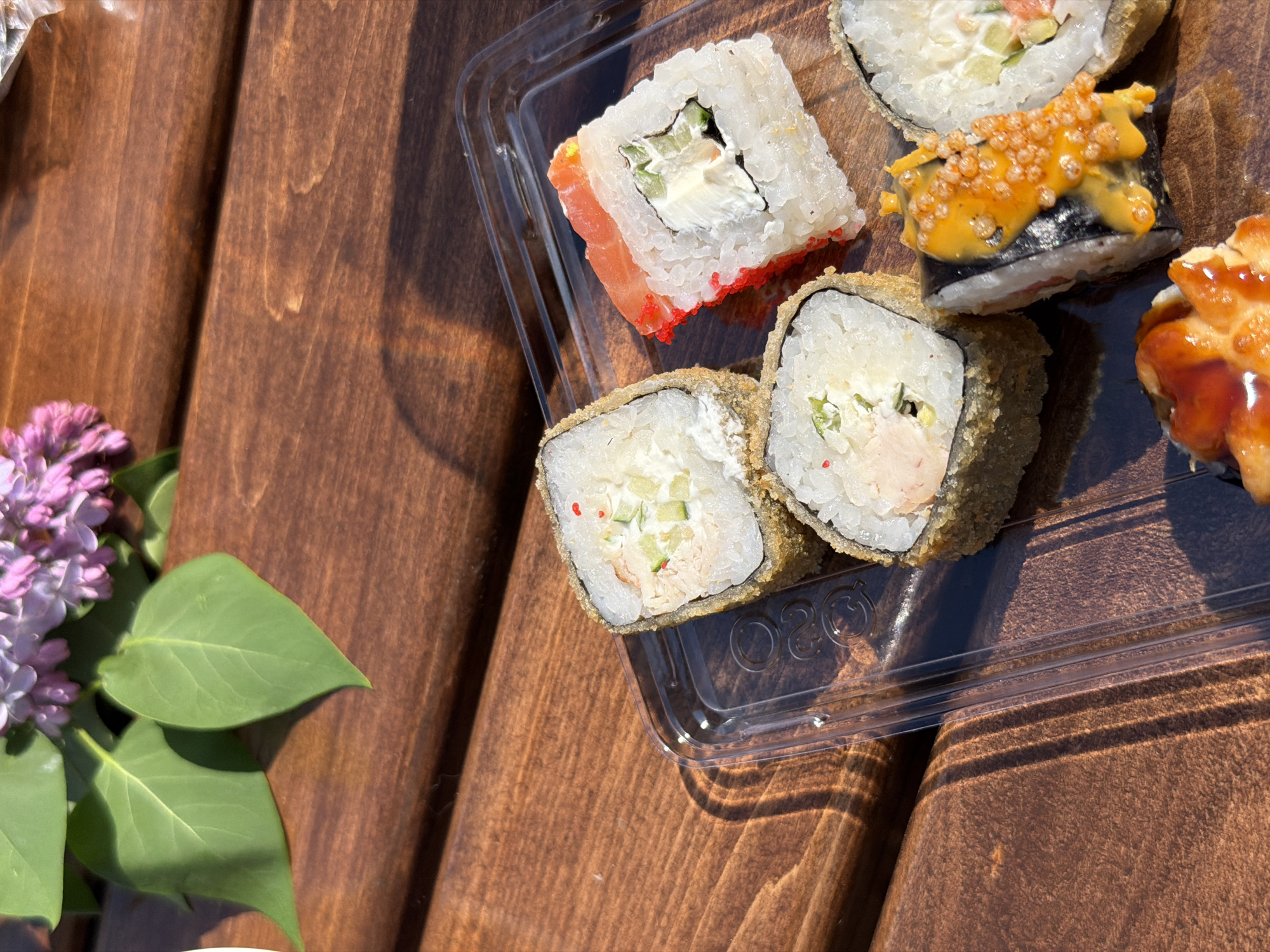 The image shows a collection of sushi rolls arranged in a clear plastic container, placed on a wooden surface. To the left of the sushi, there's a sprig of lilac blossoms. The wooden surface appears to be textured. The lighting indicates an outdoor setting, possibly a sunny day. The location is identified as the city of Chaikovsky, Russia, although there are no specific visual cues confirming this beyond the metadata associated with the image. There are no people present in the frame. The focus is on the arrangement of food and flora on the wooden surface.