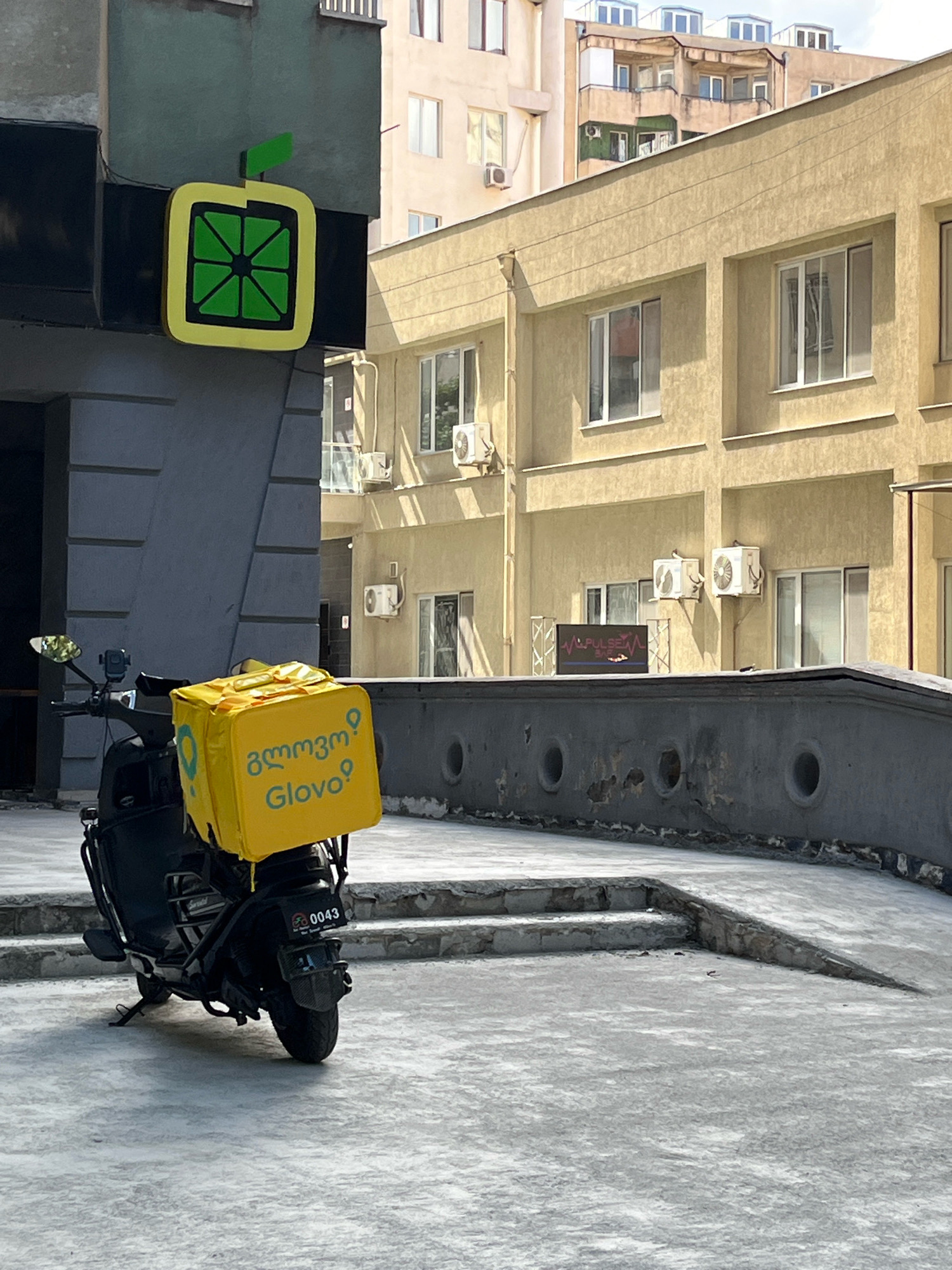 A black scooter is parked on a paved surface in front of a building. The scooter has a bright yellow insulated delivery bag on the back, which features the Glovo logo in blue. A license plate on the back of the scooter reads "0043". Above and to the left of the scooter, a sign shaped like a stylized lemon with a green stem is mounted on a building. The building behind the scooter is a light yellow, and further in the background, other buildings with visible windows and air conditioning units are present. The overall setting appears to be an urban environment.