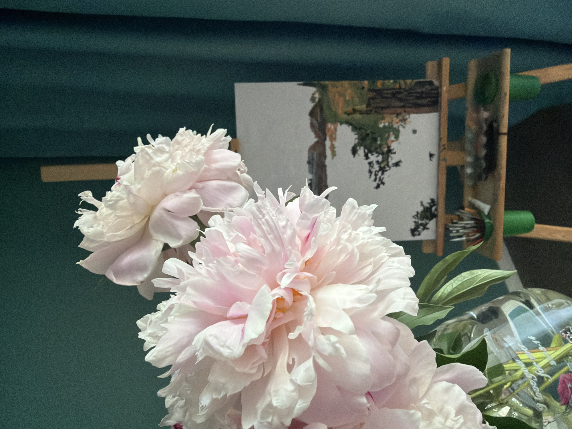 The image depicts a still life arrangement with a bouquet of pale pink peonies in a glass vase in the foreground, to the left. The vase has some inscription on it, partially visible. Behind the flowers, an easel stands with a small landscape painting on it, depicting what appears to be a body of water with buildings and trees along the shore. Various art supplies, including paintbrushes and a palette, are arranged on a tray attached to the easel. The background is a dark teal curtain. The scene is indoors, likely in a domestic setting. There are no people or animals visible. The lighting suggests it is daytime, with light coming from the side. The location is not specified within the image itself.