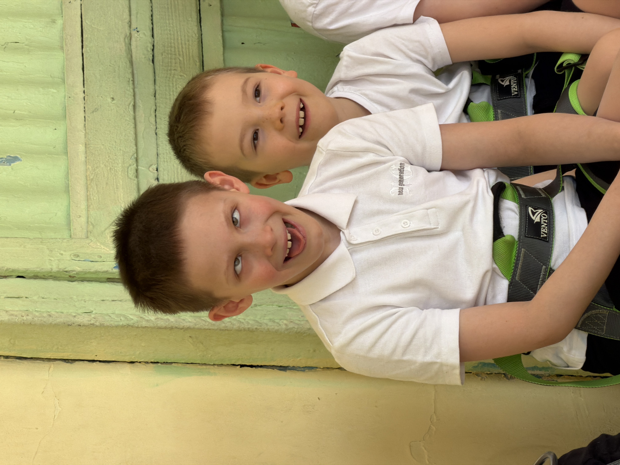 Two young boys, likely pre-teen, are captured in a bright, close-up shot. Both are wearing white polo shirts and green and black climbing harnesses, suggesting participation in an adventure activity like climbing, a ropes course, or a zip line. The harnesses clearly display the brand name "VENTO."
The boy positioned higher in the frame is smiling broadly, revealing several missing front teeth. The boy lower in the frame is also smiling widely, looking directly at the camera with a playful expression, and sticking out his tongue. His white polo shirt has "New generation" embroidered on the left chest area.
The background consists of two distinct sections. Behind the boys, there is a light green wooden wall, appearing weathered with visible vertical planks and some peeling paint. Below this, a light yellow, textured surface, possibly a concrete wall or ground, is visible. The lighting is bright, indicating it's daytime. The overall mood conveyed by the boys' expressions is happy and energetic. The location context provided is Izhevsk, Russia.