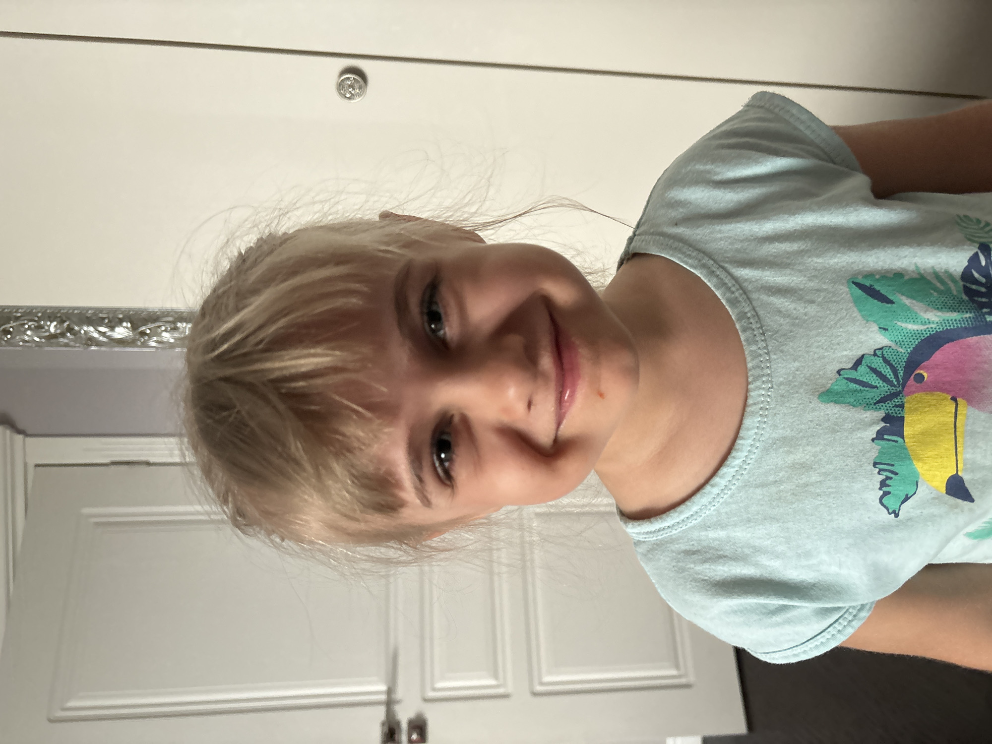 A girl with blonde hair is smiling at the camera. She is wearing a light blue t-shirt with a tropical print. The background appears to be an interior space with a white door. The location is Unknown, Unknown.