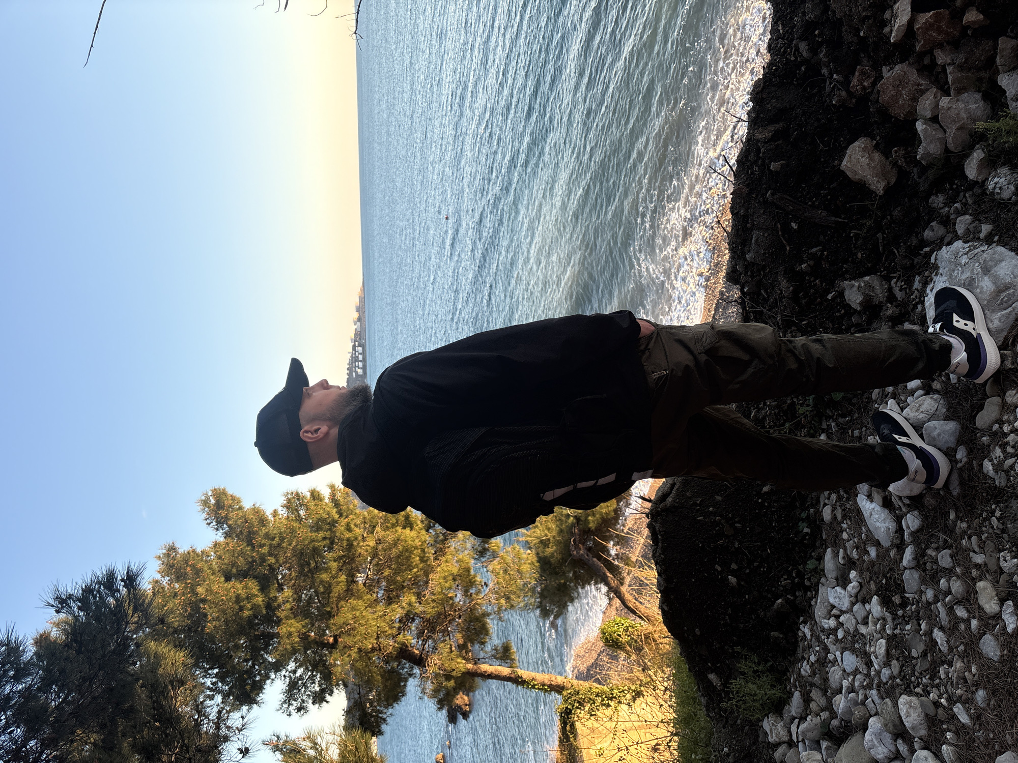 A man stands on a rocky cliff overlooking the Adriatic Sea in Bar, Montenegro. He is wearing a black jacket, dark pants, a baseball cap, and black and white sneakers. The sea is visible in the background, with trees and other vegetation in the foreground. The sky is clear and blue.