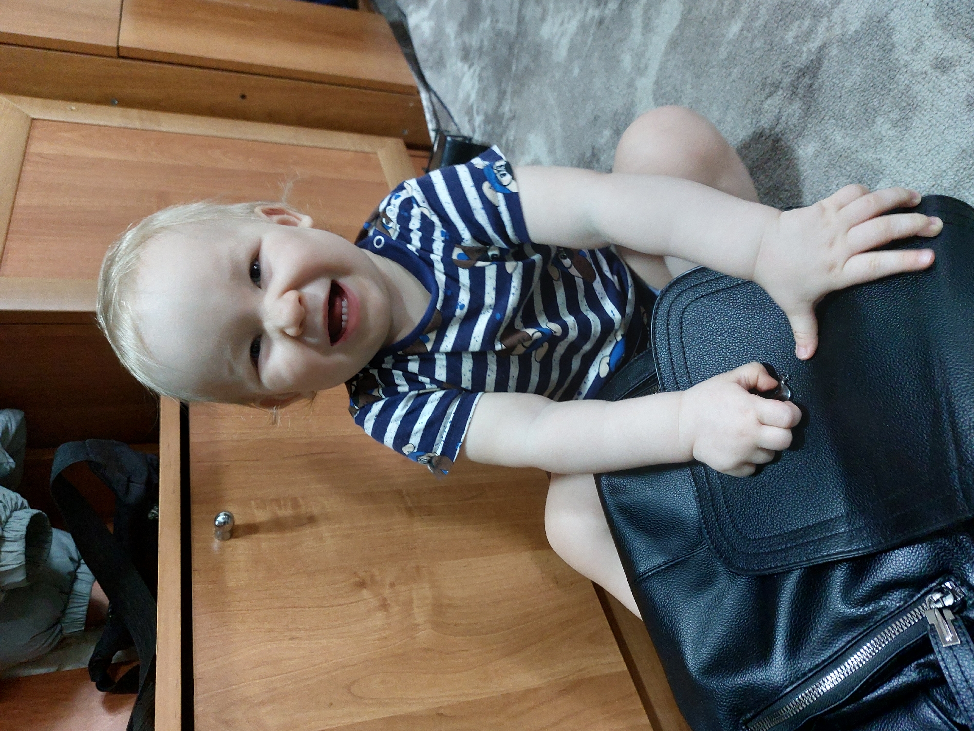 A young child with blonde hair, wearing a blue and white striped shirt with cartoon bear images, is seated on the floor next to a wooden cabinet. The child is smiling and appears to be reaching for or playing with a black leather bag. The background includes a carpeted area.