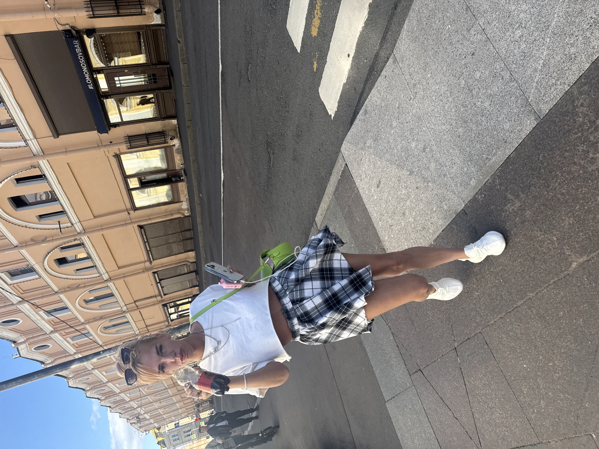 A blonde woman wearing a white crop top, a plaid skirt, and white sneakers stands on a sidewalk in Saint Petersburg, Russia. She holds a bottle of soda in her right hand and a smartphone in her left. A green purse with a strap is slung over her shoulder. In the background, a building with beige walls and arched windows stands next to a street with a crosswalk. A sign on the building reads "#LOMONOSOVBAR".