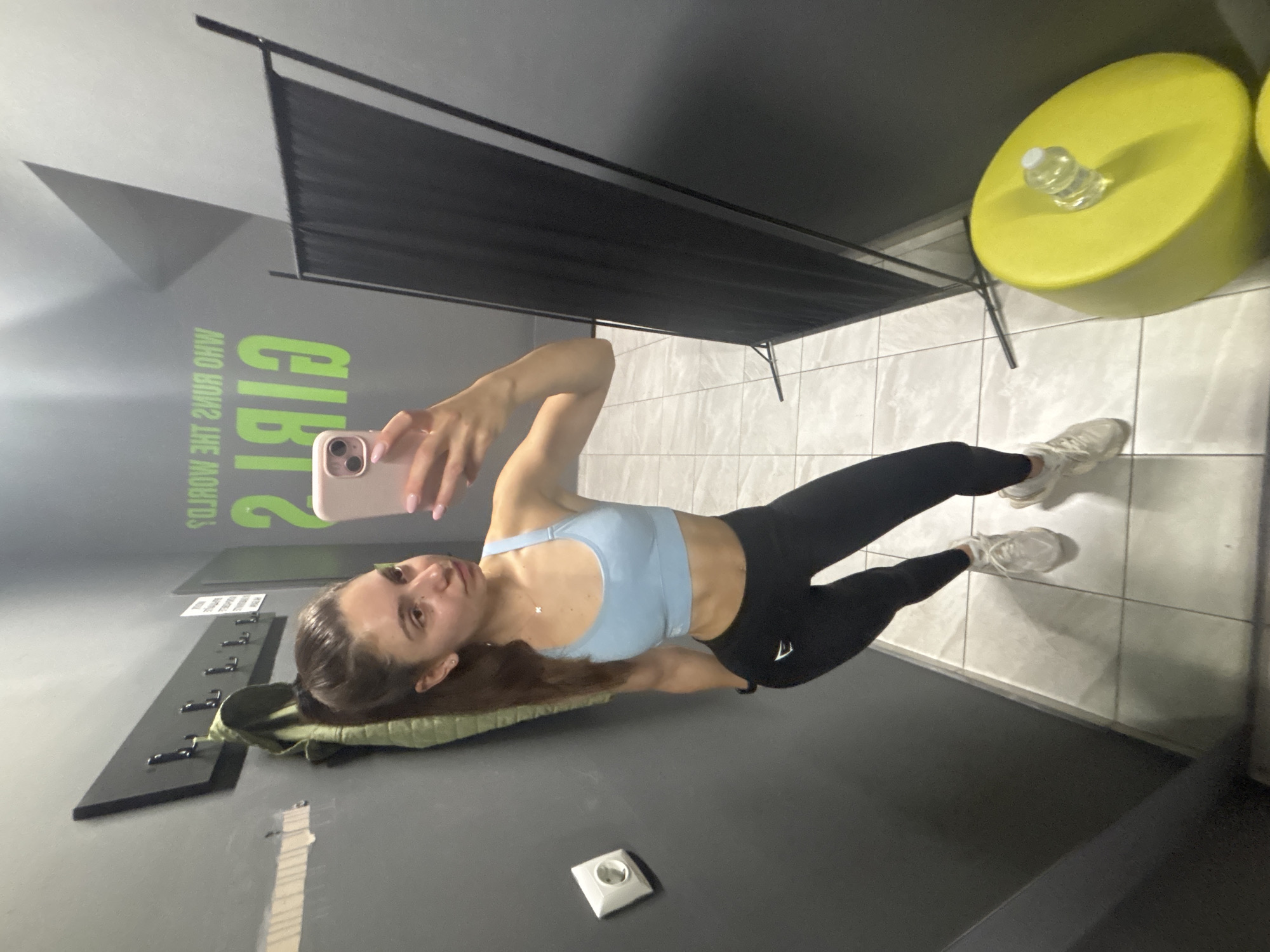 A young woman with brown hair in a ponytail, wearing a light blue sports bra and black leggings, stands in a gym locker room, taking a mirror selfie with her pink smartphone. She is wearing white athletic shoes. Behind her, a green graphic on the wall reads "GIRLS WHO RUN THE WORLD". A coat rack with a green jacket hangs on the wall to her left, and a power outlet is visible. To her right, a black folding screen stands, and a bright yellow cylindrical object with a clear lid is on the floor. The floor is tiled with large, light grey squares. The lighting is artificial and somewhat dim.