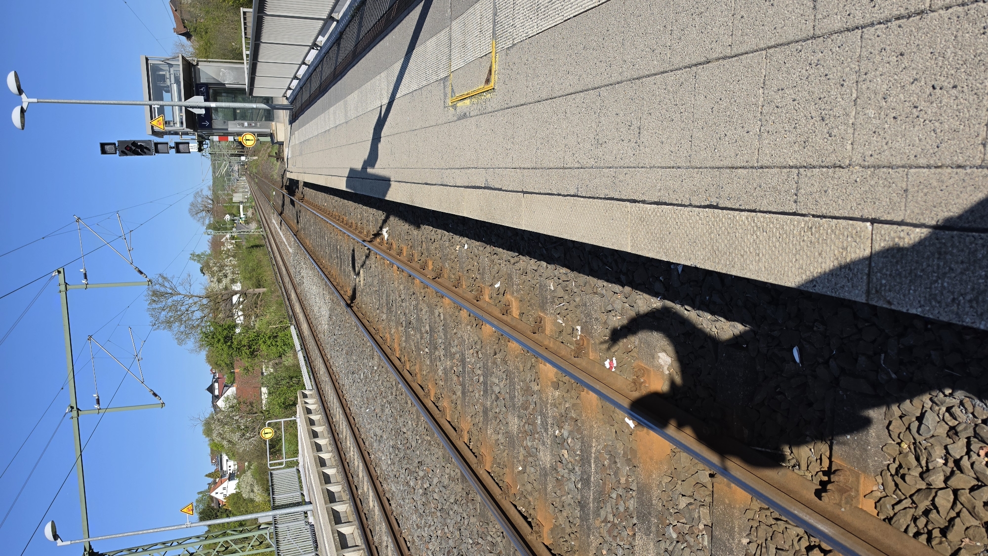 The image, captured from a slightly elevated perspective and rotated 90 degrees clockwise, presents a bright, sunny view of a train platform alongside railway tracks. The concrete platform dominates the upper half of the frame, extending into the distance. It features various textures, including smooth and textured concrete slabs.
On the platform, a silver lamppost with two upward-facing lights is visible towards the foreground. Further along, a pole supports railway signals displaying multiple red lights, indicating a stop or caution. Adjacent to this, a modern-looking, glass-enclosed structure, possibly an elevator or waiting shelter, stands. Several yellow warning signs are affixed to nearby structures: a triangular sign with a black exclamation mark, a circular sign with a capital 'I' (likely indicating information or a lift), and another sign featuring an arrow pointing right. A yellow, U-shaped fixture is embedded in the platform near the edge.
Two sets of railway tracks run parallel in the lower half of the image, with rusty rails resting on grey gravel ballast. Above the tracks, an extensive overhead electrical (catenary) system is supported by metal poles and gantries, typical of electrified rail lines. A striking, dark shadow resembling a large bird, possibly a swan with its curved neck, stretches across the tracks and onto the platform from the bottom-right corner, a distinctive feature.
In the background, beyond the railway infrastructure, residential houses with tiled roofs are visible amongst lush green trees and some white-flowering trees, suggesting a springtime setting. The sky is a clear, vibrant blue, indicating fair weather. The long, distinct shadows on the tracks and platform suggest it is either morning or late afternoon. The scene is devoid of people or trains, offering a quiet, static view of the railway environment.