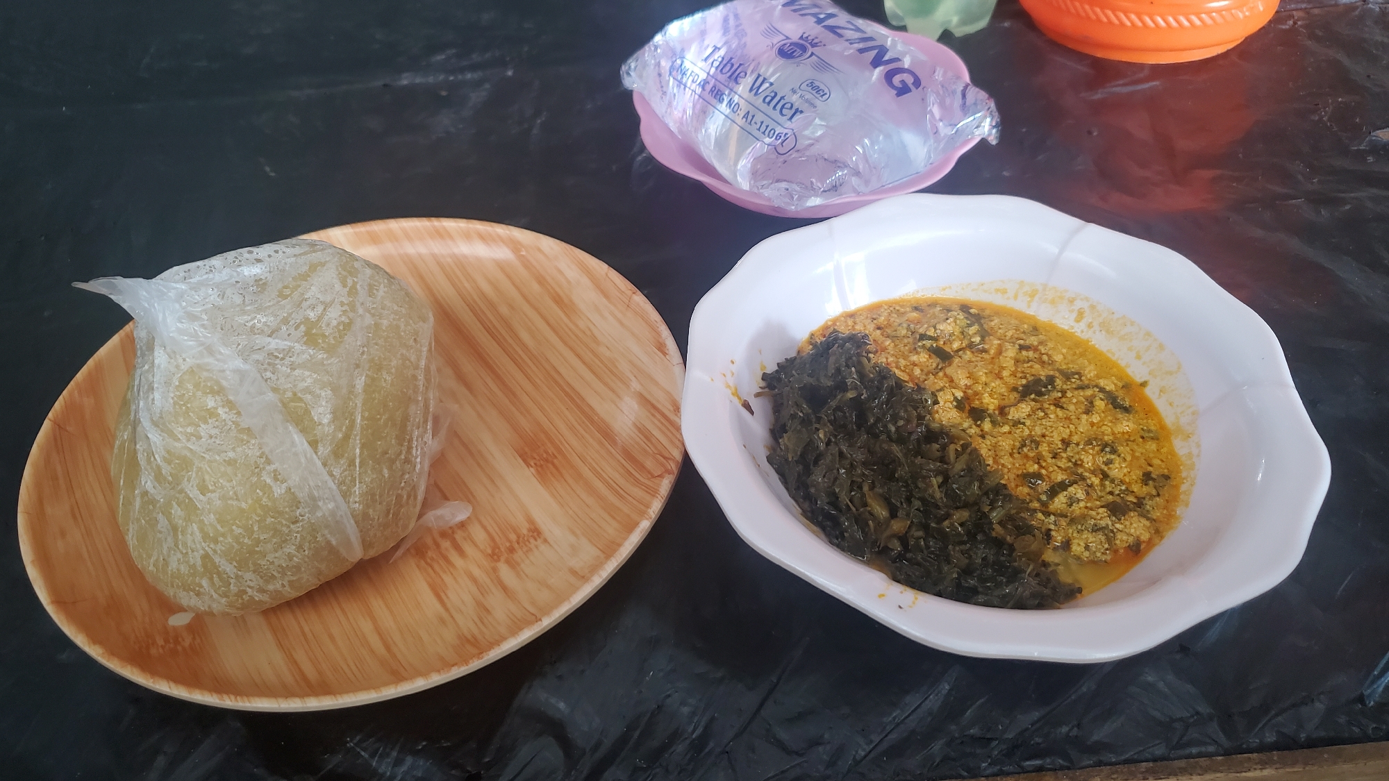 A light meal is presented on a dark, possibly plastic, tablecloth. On the left, a light brown wooden plate holds a portion of Fufu, a staple food in West Africa, wrapped in clear plastic. To the right, a white, scalloped-edge bowl contains a dish composed of two distinct sections: a dark green, leafy vegetable on the left, and a yellowish-orange mixture on the right, which appears to be a sauce with small pieces of food, possibly scrambled eggs or ground nuts, mixed in. In the background, a pinkish-white plastic dish holds a sealed packet of "AMAZING Table Water," with text indicating it is 50cl. An orange, ribbed container is partially visible to the far right. The lighting suggests an indoor setting, likely a home, with a diffused light source. The image is taken from a top-down perspective. No individuals are visible in the image. The setting is consistent with a casual dining experience in Jalingo, Nigeria.