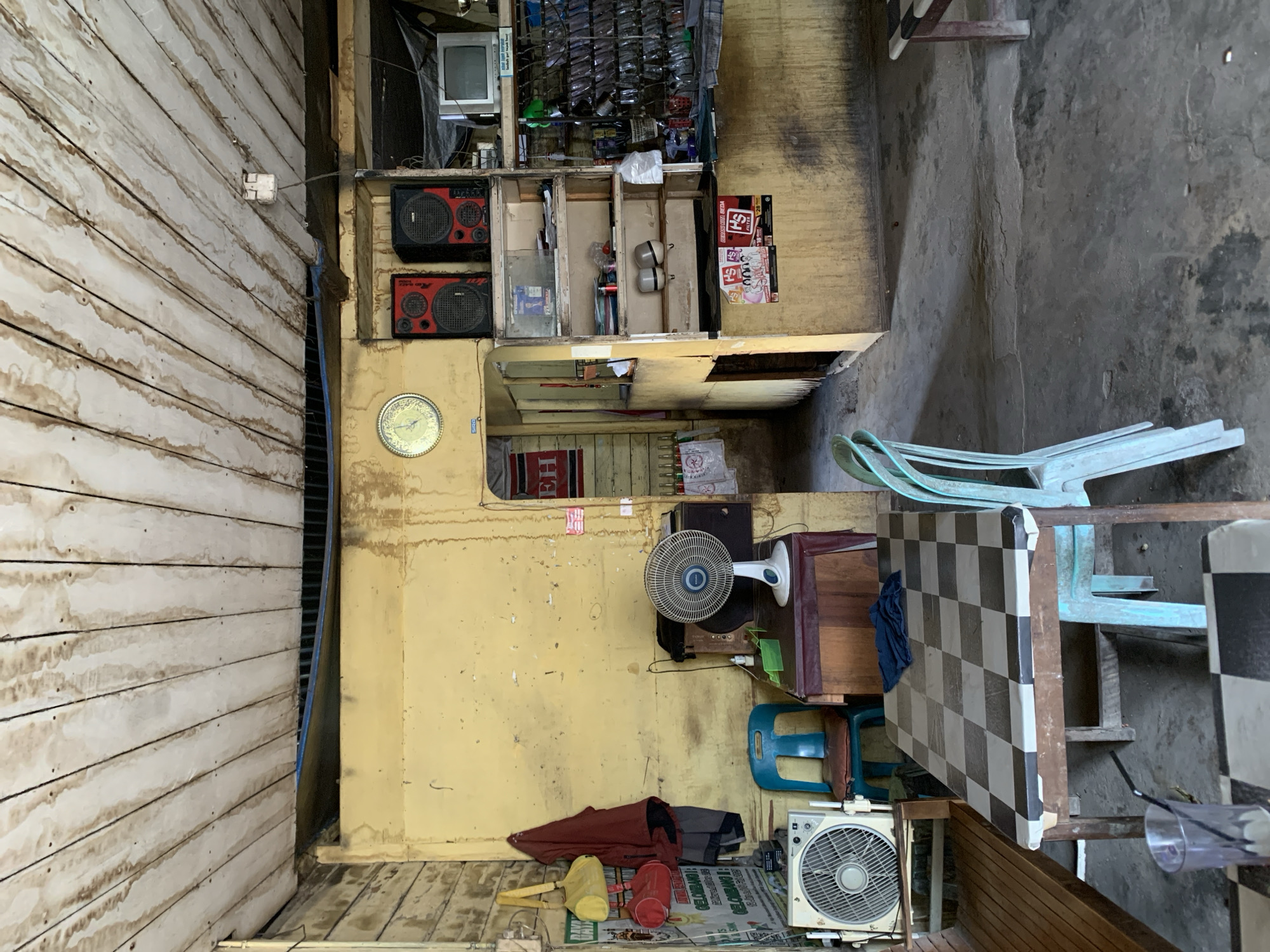 This image depicts an interior scene of what appears to be a small shop or eatery in Meunasahdrang, Indonesia. The room is aged and shows signs of wear. The ceiling is constructed of weathered wooden planks with dark stains. The walls are painted a faded yellow and have numerous smudges and water stains.

In the foreground, a table with a checkered black and white surface is set with a glass containing a dark beverage and a straw. A blue cloth is draped on the table. Two light blue plastic chairs are placed around the table, one of which is partially obscured.

To the left of the table, a white box fan sits on a wooden bench. Next to the fan, a bright blue plastic chair is visible. Behind the fan and chair, a dark wooden cabinet is present.

In the mid-ground, a white oscillating fan is positioned on top of the dark wooden cabinet. Further back, a doorway reveals a glimpse of another area with red and white striped walls. Above the doorway, a circular clock with a gold frame is mounted on the yellow wall, beside two red and black speakers. To the right of the speakers, a small, older television set is visible on a shelf.

A shelving unit to the right of the speakers displays various items, including what appear to be bags or packaged goods on the lowest shelf, and some containers on the shelf above. Faded posters are affixed to the wall below the shelves. The floor is concrete and shows some discoloration and cracks. The overall impression is one of a well-used, humble establishment.
