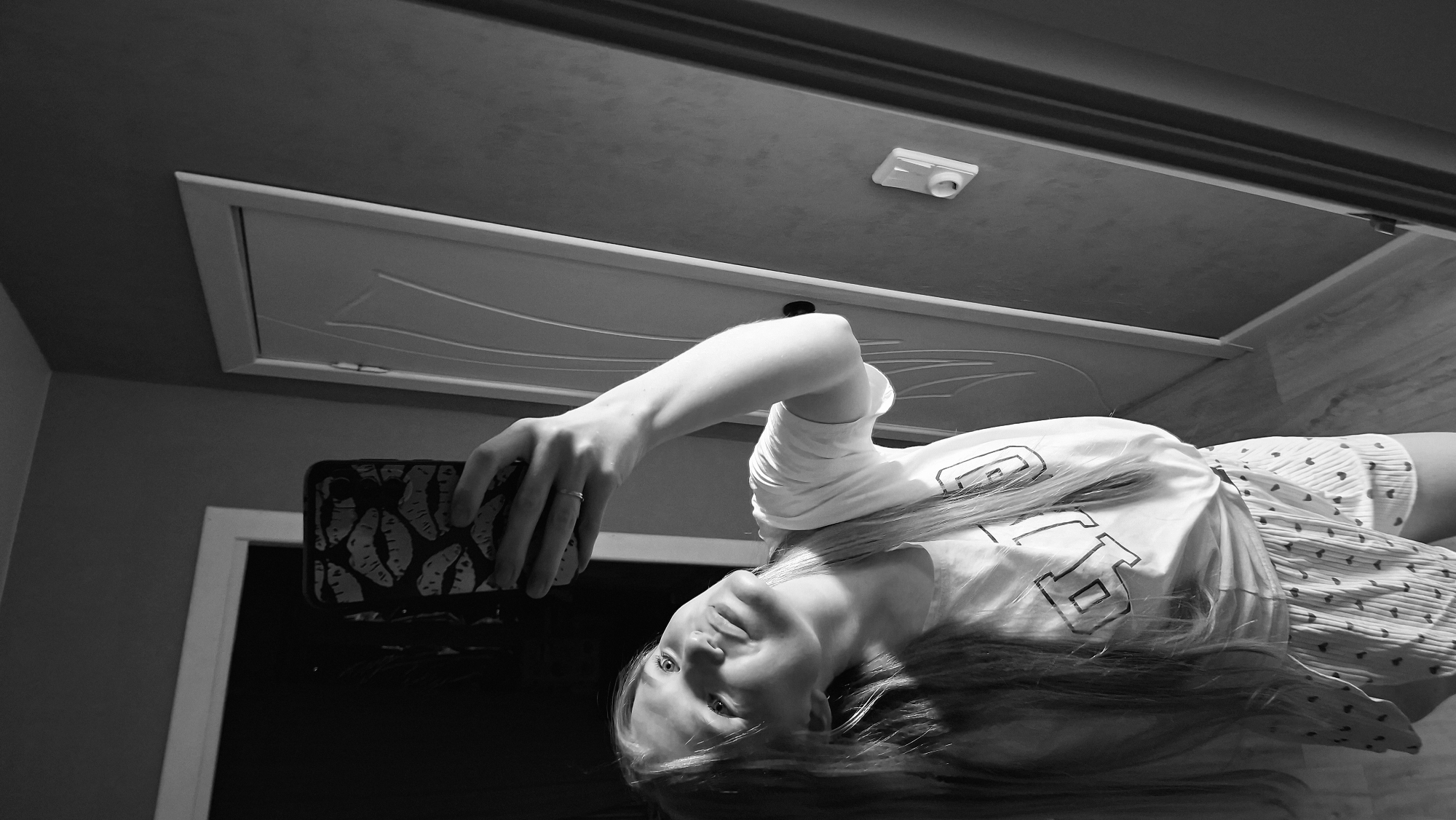 In a black and white, low-light photograph, a young woman is captured taking a mirror selfie in what appears to be a bedroom or dressing area. She is standing in front of a door and a wall with some light fixtures. The woman has long blonde hair and is wearing a t-shirt with a graphic design and patterned shorts. She is holding up a smartphone with a distinctive case patterned with lips. Her arm is raised to take the photo, and her face is turned upwards, looking at her reflection. The room itself is sparsely furnished, with a doorway and wall visible in the background. The overall impression is casual and intimate. The scene is suggestive of domestic personal space within the city of Chaikovsky, Russia.