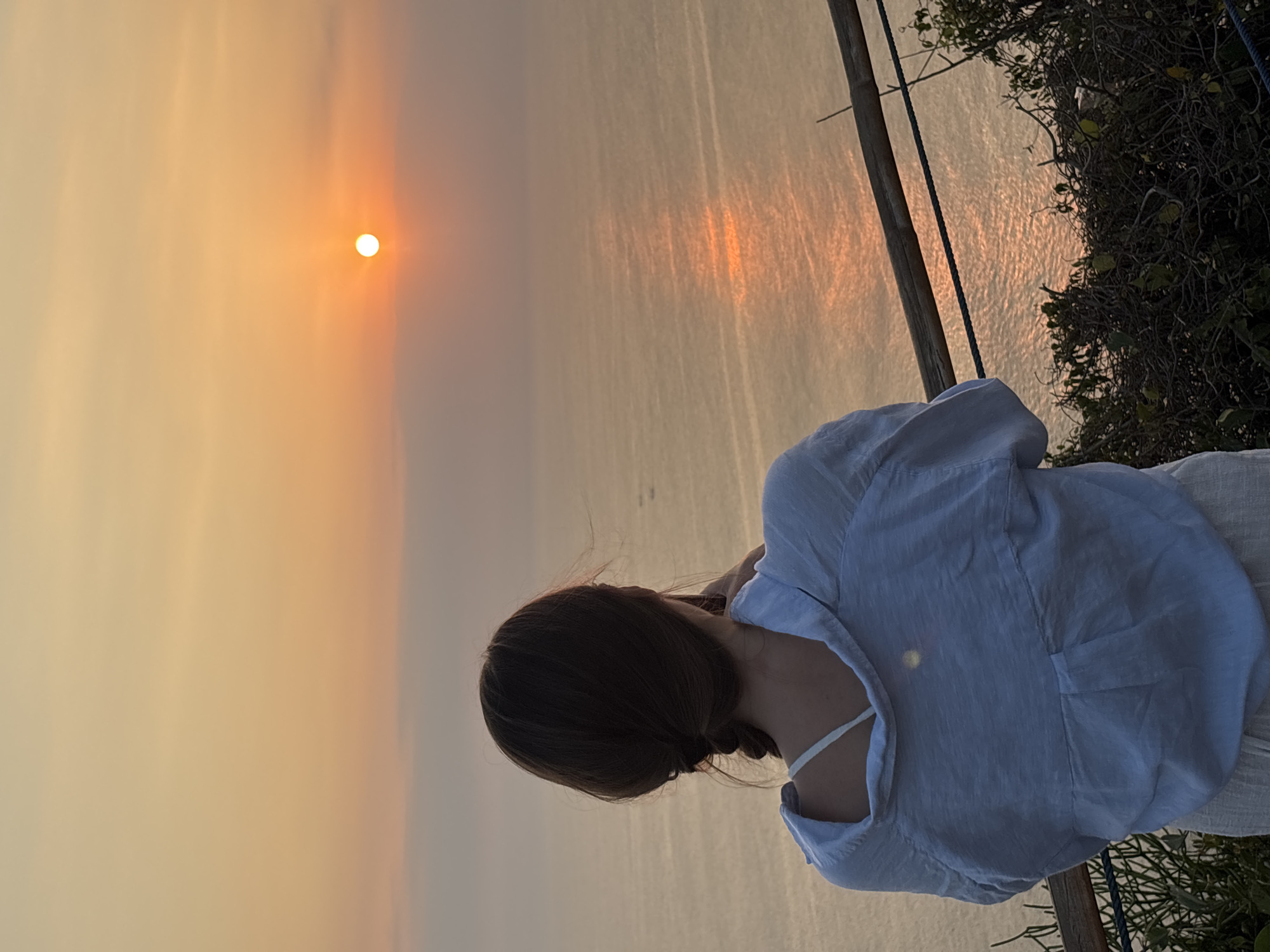 A woman, viewed from behind, stands on an elevated vantage point overlooking a calm ocean during sunset. The sun, a bright orb, is low on the horizon, casting a warm orange glow across the sky and reflecting on the water's surface. The woman has dark hair styled in a braid and wears a light-colored, loose-fitting shirt and a white bra. She appears to be holding a camera or phone up to take a picture. In the foreground, a wooden railing and lush green vegetation frame the scene. The location is Kuta Selatan, Indonesia, known for its scenic coastal views and sunset vistas.