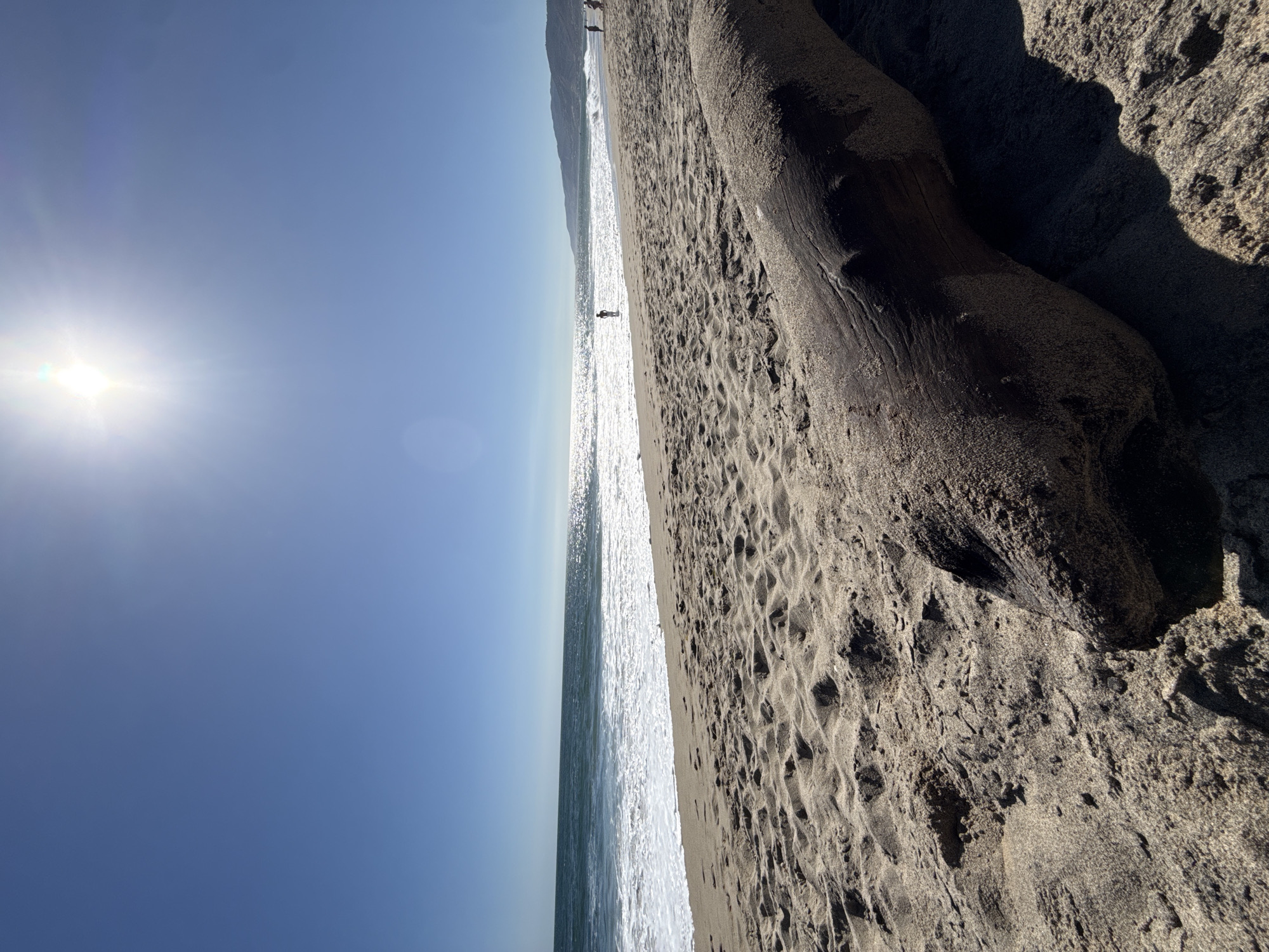 A low-angle, wide shot captures a sun-drenched beach in Malibu, California. The foreground is dominated by a weathered, dark brown driftwood log lying on the sand. The sand itself is light brown and textured with numerous small mounds and depressions. The ocean is visible in the mid-ground, with blue waves breaking onto the shore and white foam receding. The sun, a bright white orb with a visible lens flare, shines intensely in the upper center of the frame, casting a glare on the water. The sky is a clear, deep blue. In the distance, the coastline curves, with hills covered in vegetation forming a backdrop. Several small human figures are present on the beach and in the water: one person stands in the surf, and a few others are walking along the shoreline further down the beach. The overall impression is one of a peaceful, sunny day at the beach.