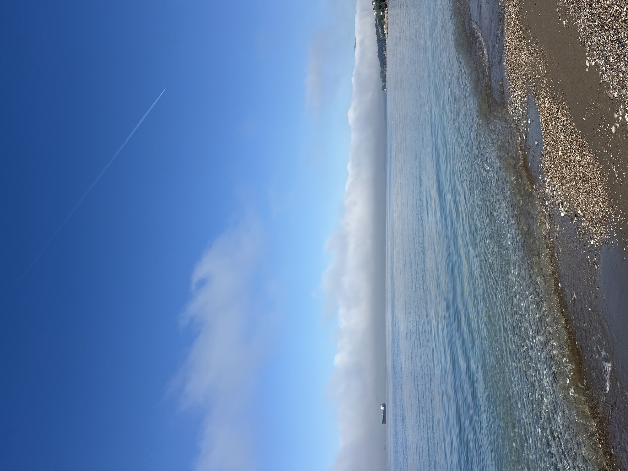 The image depicts a shoreline scene in Šušanj, Montenegro. A body of water, likely the Adriatic Sea, meets a beach composed of dark sand and small shells. Gentle waves lap the shore. The sky is partially clear, with a contrail visible in the upper left. Clouds obscure the distant coastline. A cargo ship is visible near the horizon. The image is rotated 90 degrees counter-clockwise.
