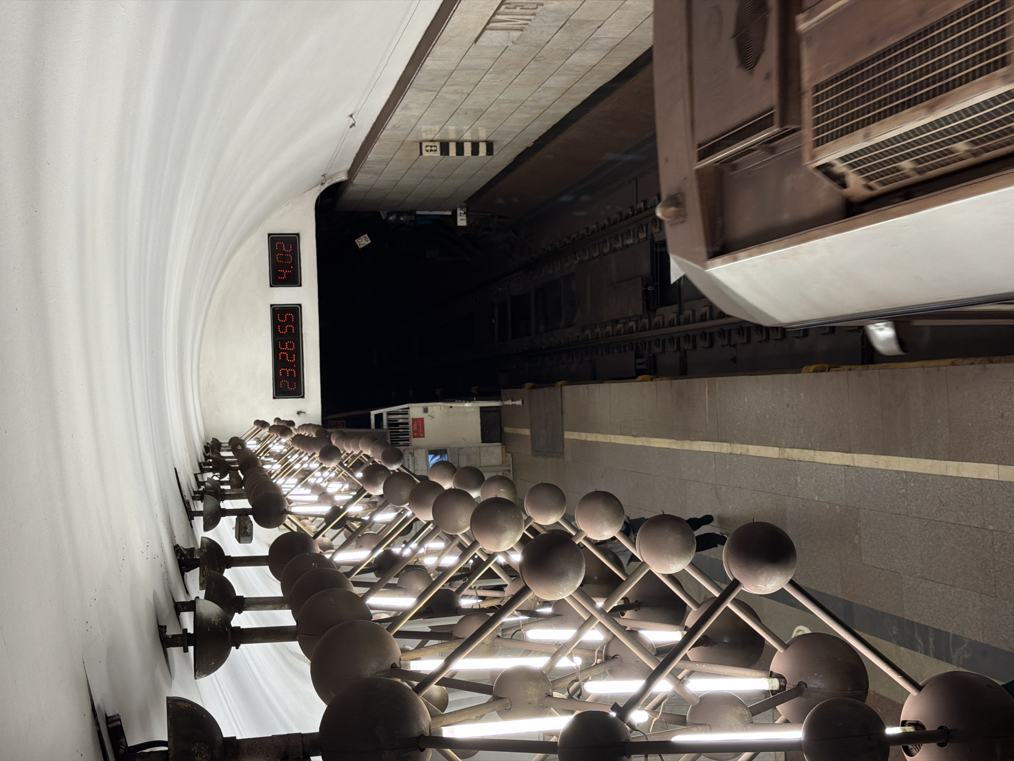 This photo shows the Moscow Metro station Elektrozavodskaya. The picture looks sideways because it was taken in portrait format, and not reoriented to a landscape aspect ratio.
The primary focus of the image is a decorative lighting structure consisting of numerous spherical lamps arranged in a grid pattern along the station's platform. These lights are a distinctive feature of the station's design.
To the right of the lighting fixtures, we see the station platform with passengers waiting along the edge. A train is arriving or departing, with a portion of a rail car visible.
The walls of the station are white and curved, with digital clocks displaying the time (23:26:55 and 4:02). There is also an exit with a black-and-white square design. The photo captures the typical environment of a bustling metro station.