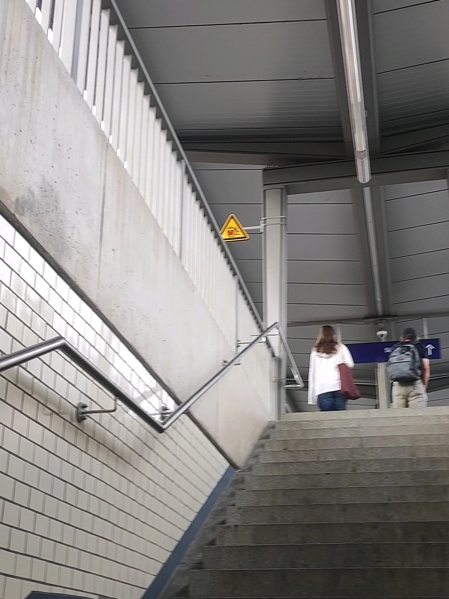 The image shows an outdoor staircase at the Nuremberg subway station. Two individuals are walking up the stairs, one in front of the other. The person in front is wearing a white shirt and jeans, carrying a dark red bag. The person behind is wearing a backpack and khaki shorts. The staircase is made of concrete, with a metal railing on the left side. The walls on the left are tiled with white rectangular tiles. Above the staircase, there is a grey, corrugated metal ceiling. A yellow triangular warning sign with black symbols is visible on a metal pole to the left. A blue sign with white text and an arrow is mounted on a pole further up the stairs, indicating a direction. The lighting suggests it is daytime.
