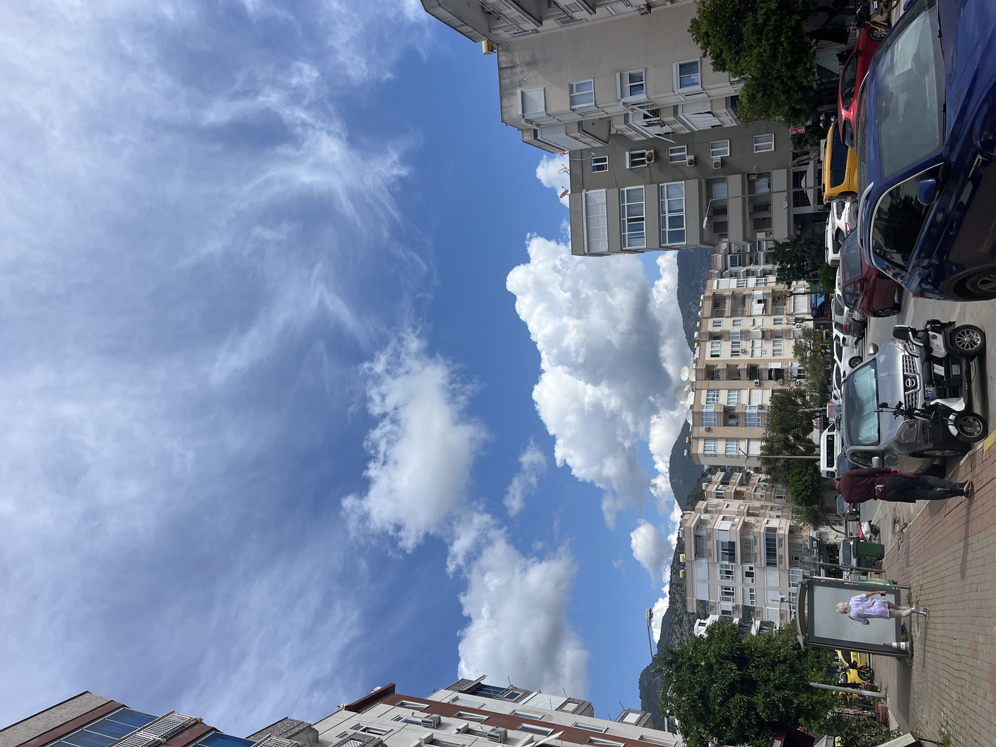 The image shows a street scene in Alanya, Turkey, with buildings, parked vehicles, and people. The buildings appear to be residential apartments. Various parked cars and motorbikes line the street. A few people are walking on the sidewalk, including an elderly person standing near an advertisement board. Above the buildings is a blue sky with scattered clouds. Mountains can be seen in the background.