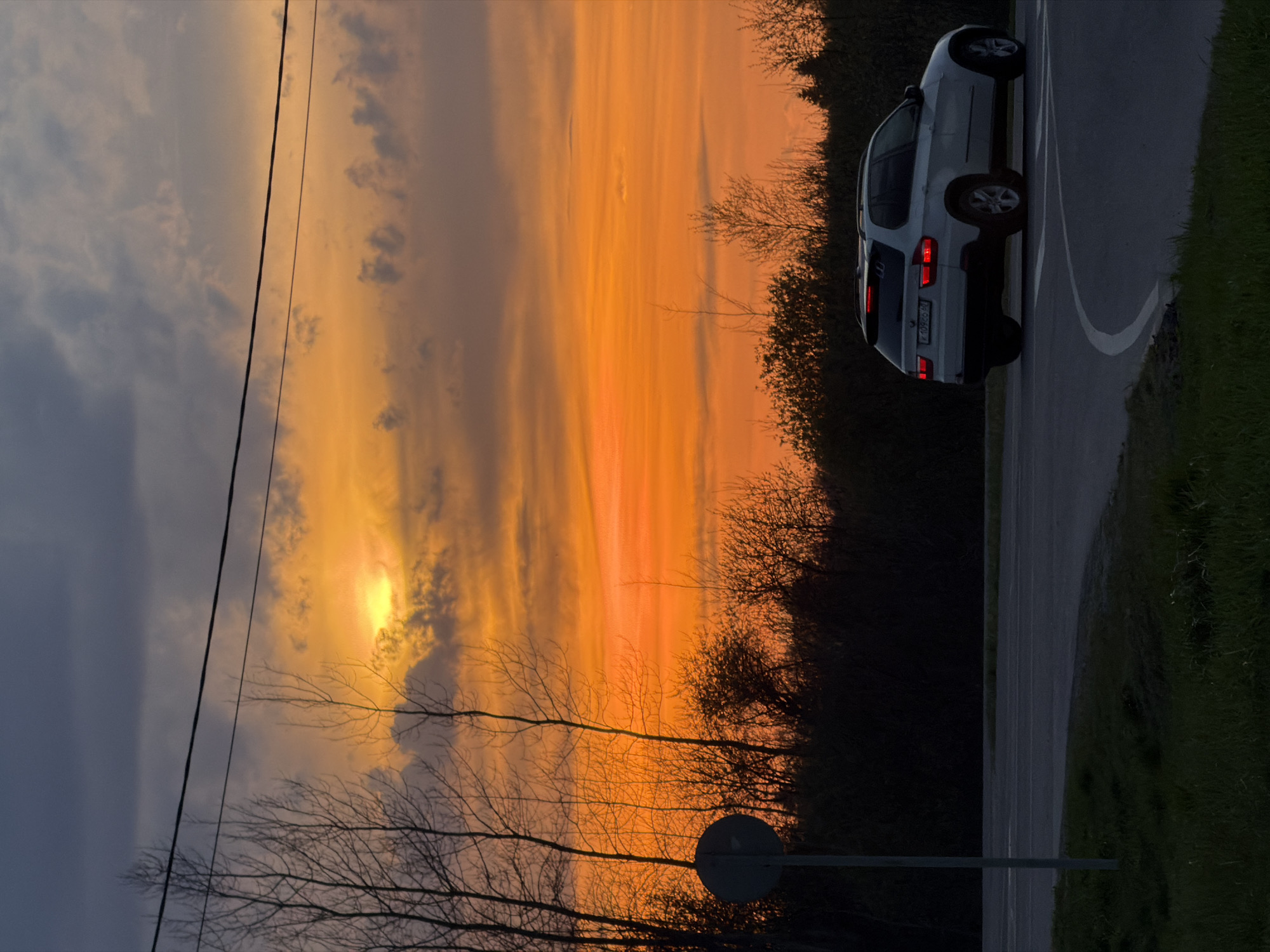 The image depicts a silver-colored BMW X3 SUV parked on the right side of a curved road, likely in Ostrov, Russia, as indicated by the license plate “K709EO60”. The car's rear lights are illuminated. The setting is a rural or suburban area with dense foliage. In the background, a striking sunset dominates the sky, with shades of orange and yellow filtering through the clouds. Power lines are visible against the left side of the sky, which is dark. The road is gray, and green grass lines its edge. A street sign is partially visible.