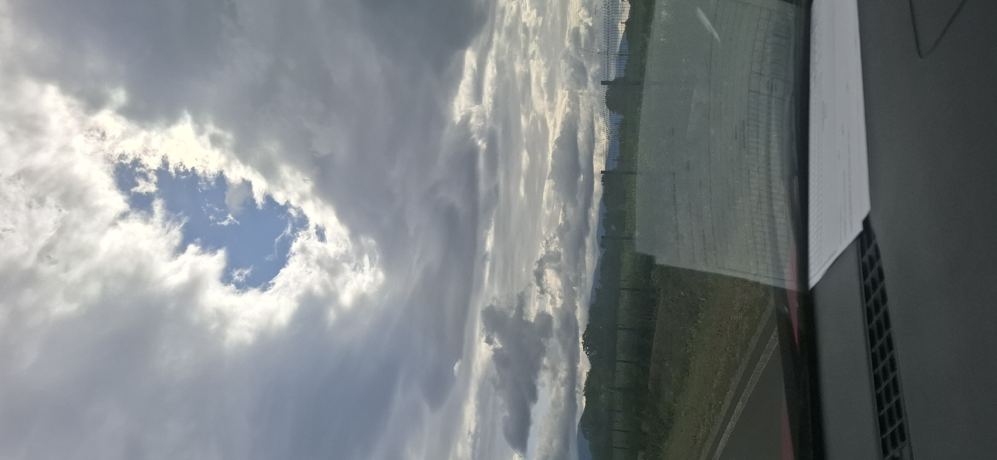 This image shows a view from inside a vehicle, looking out through the windshield. The foreground of the image shows the dashboard of the car, with a piece of paper resting on it. The windshield is visible, reflecting the sky and the landscape outside.
The sky is predominantly filled with dark, heavy clouds, with a bright patch of blue sky visible in the upper center, peeking through a gap in the clouds. This opening in the clouds is illuminated by sunlight, creating a contrast with the surrounding darker clouds.
Beyond the windshield, a fence is visible, made of metal posts and wire mesh. Behind the fence, there is a green field with some vegetation and trees. In the distance, rolling hills or mountains are visible, partially obscured by the clouds and haze. The overall atmosphere suggests an overcast or partly cloudy day, possibly before or after rain. The scene is devoid of people or any specific action. The location in the city of Dolno Sahrane, Bulgaria, cannot be determined from the visual information present in the image.