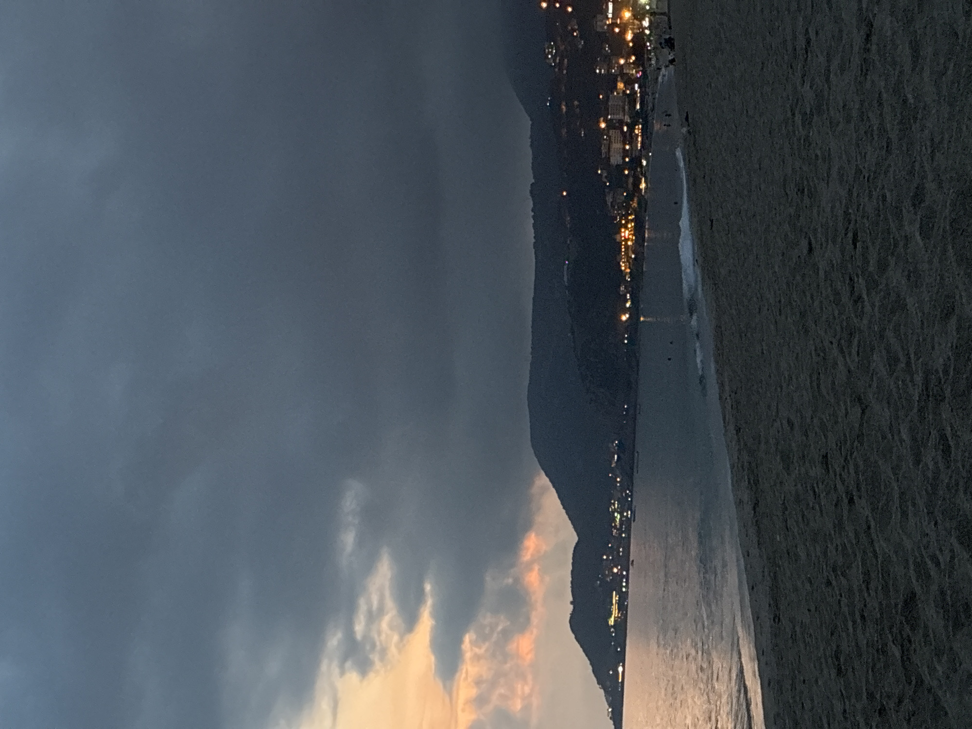 A dusky, atmospheric view of Alanya, Turkey, at twilight. The foreground features a sandy beach with subtle textures. Beyond the beach, the calm sea meets the shore, with gentle waves. Along the coastline, a densely populated town sprawls across the foothills of dark, silhouetted mountains. Buildings are illuminated with warm, artificial lights, creating a twinkling effect against the darkening landscape. The sky above is heavy with dramatic, dark clouds, but a sliver of the horizon to the left shows a hint of the setting sun, casting a faint orange and pink glow through the clouds. Several small boats are visible on the water, and a few distant figures can be seen on the beach, suggesting a serene evening by the sea.