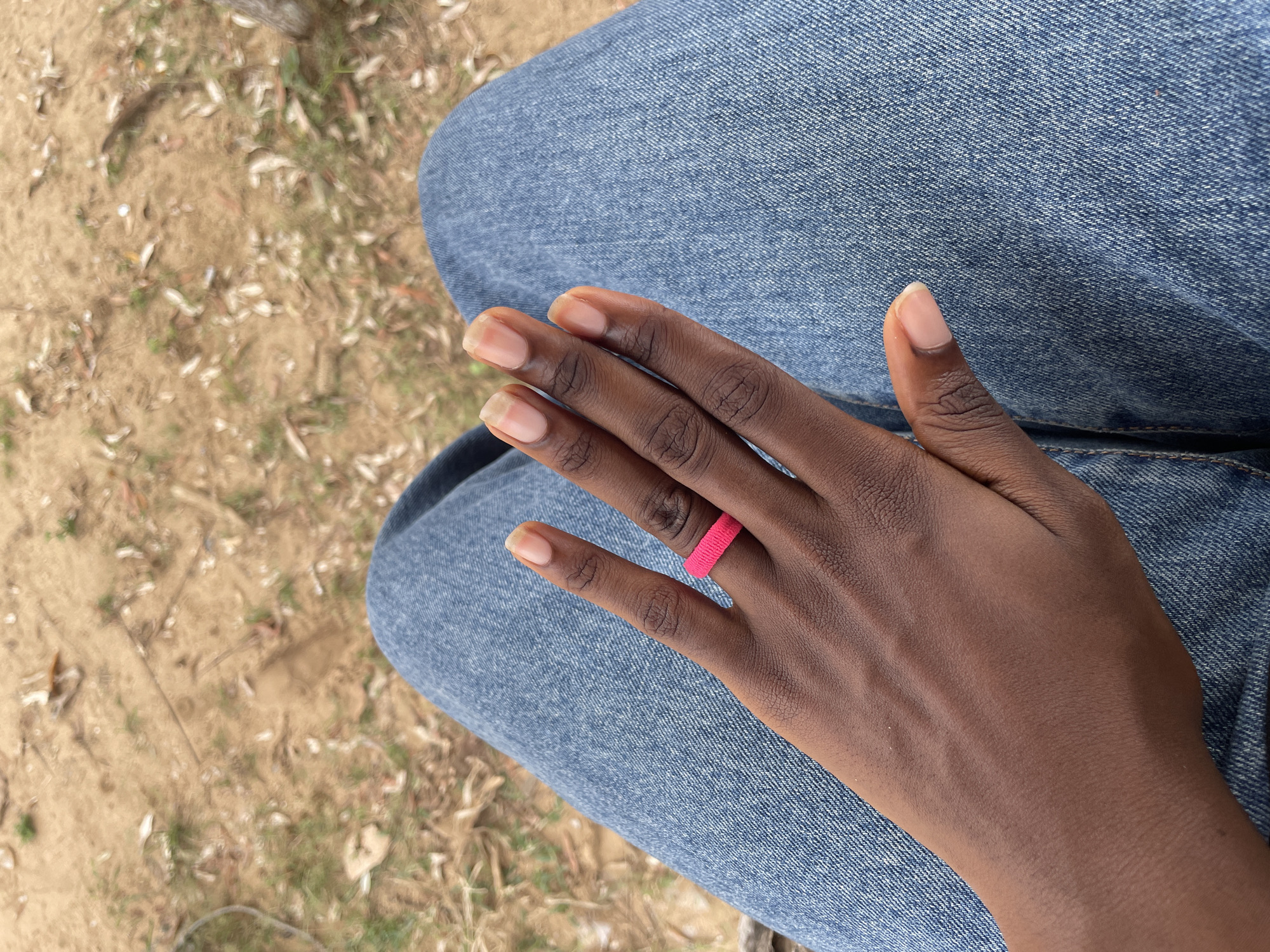 A close-up overhead shot shows a person's left hand, adorned with a bright pink ring, resting on their lap, which is covered by blue denim jeans. The background consists of dry, sandy earth scattered with fallen leaves and patches of grass, suggesting an outdoor environment. The image was taken in the city of Iyafin, Nigeria.
