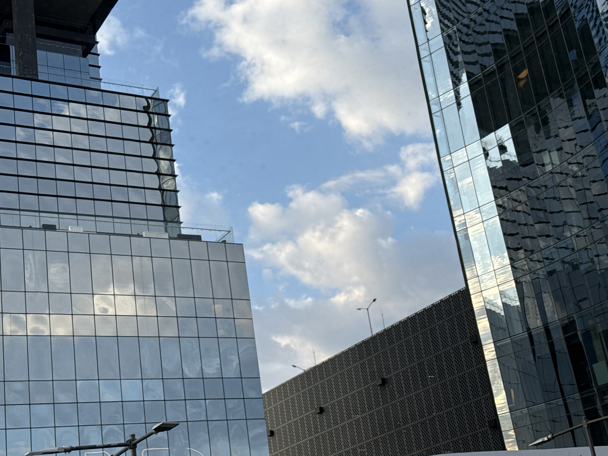The image shows a low-angle view of buildings in the city of Puebla, Mexico. Two modern high-rise buildings dominate the scene, reflecting the sky with scattered clouds. One building features a grid-like facade, while the other displays a more complex, irregular surface pattern. Between the high-rise buildings, a third, shorter building with a dark, perforated facade can be seen. A street lamp is visible in the foreground.