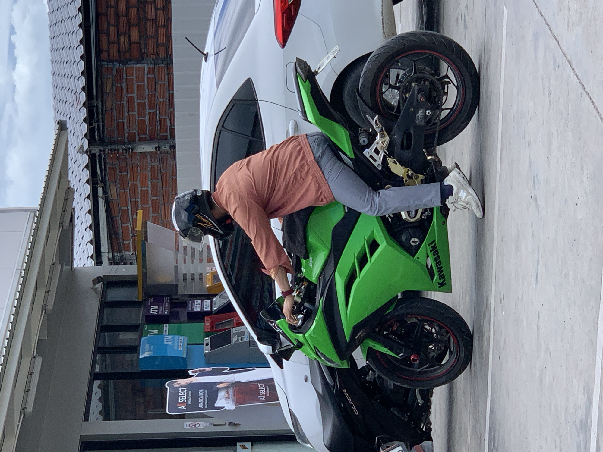 An outdoor scene captures a person seated on a bright green Kawasaki sport motorcycle, stationary on grey concrete pavement with white lines. The individual wears a black full-face helmet, a salmon-orange long-sleeved shirt, grey trousers, and white New Balance sneakers, with their right foot touching the ground. The motorcycle features black wheels with red trim and gold-colored footpeg components.
To the left of the green motorcycle, a dark Honda Click scooter is partially visible. Behind the motorcycle, a white sedan is parked. The background features a commercial building with a glass storefront, revealing three distinct ATMs: blue, green, and purple, all marked with "ATM." An "A SELECT" advertising stand to the left of the storefront promotes "SPECIALTY COFFEE" and "ARABICA 100%," featuring a male model. A no-smoking sign is also visible near the stand. The building includes a flat grey canopy with exposed fluorescent lights and a sloped, light-colored tiled roof. Beyond the roof, a red brick wall is partially visible beneath an overcast sky. While the context is consistent with Thailand, the specific location of Baan Bo Phut cannot be definitively determined from the visual evidence.