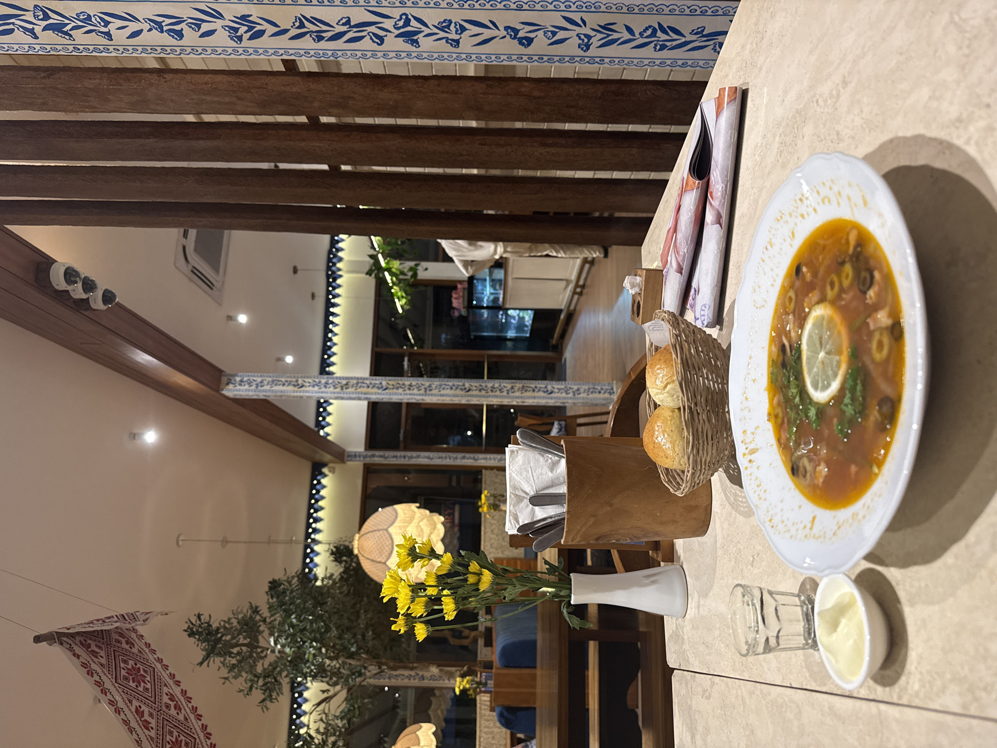 The image depicts a table setting in what appears to be a restaurant in North Kuta, Indonesia. A plate of soup garnished with a lemon slice and herbs dominates the right side of the frame. Next to it are small bowls or glasses, one containing a white substance that could be cream or sauce. A basket of rolls sits behind the plate, alongside a wooden utensil holder filled with silverware and napkins. A white vase containing yellow flowers is positioned in front of the basket. In the background, the restaurant's interior is visible, featuring wooden beams, patterned ceilings, hanging decorations, plants, and tables with blue seating. The overall ambiance suggests a dining experience in a well-decorated and inviting establishment.