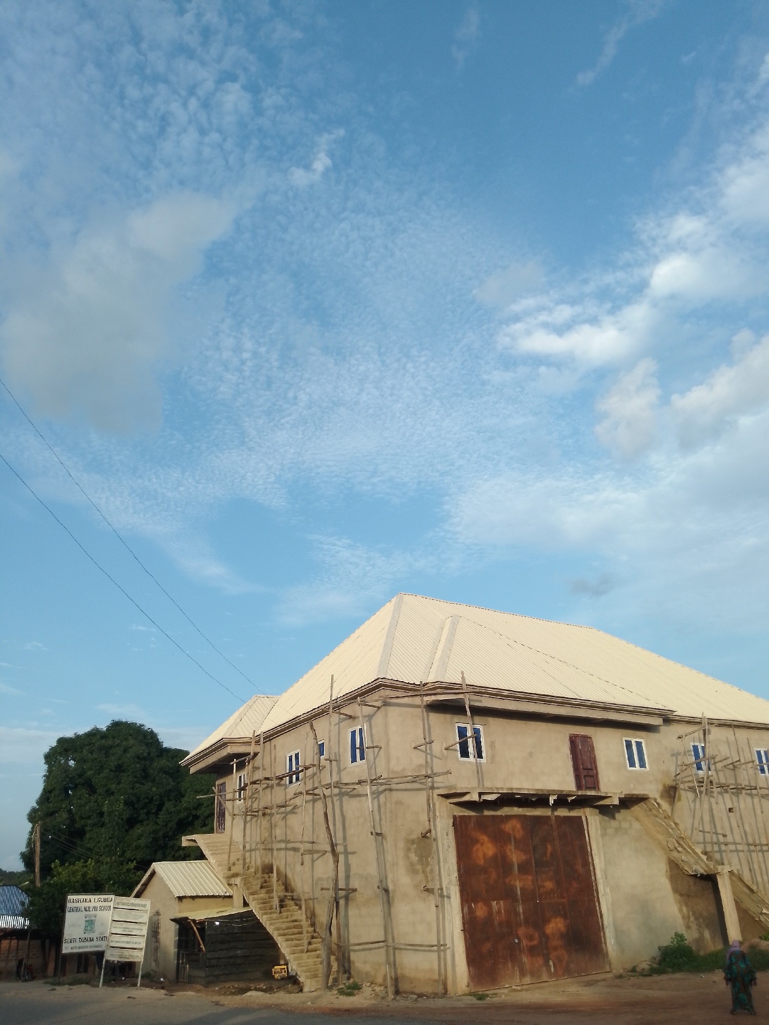 The image depicts the construction site of Gashaka LGA Central Nur/Pry School in Serti, Nigeria. A large, unfinished building with a light-colored corrugated metal roof dominates the scene. Scaffolding made of wood is attached to the exterior walls. Several windows with blue frames are visible on the upper floors. A large, rusty metal door is situated on the ground floor. An external staircase leads to an upper level of the building. To the left of the main building, a smaller structure with a similar roof is partially visible. A signpost in the foreground provides information about the school. A large, green tree stands behind the buildings. The sky above is blue with scattered white clouds. Two individuals are present in the lower right corner of the image.
