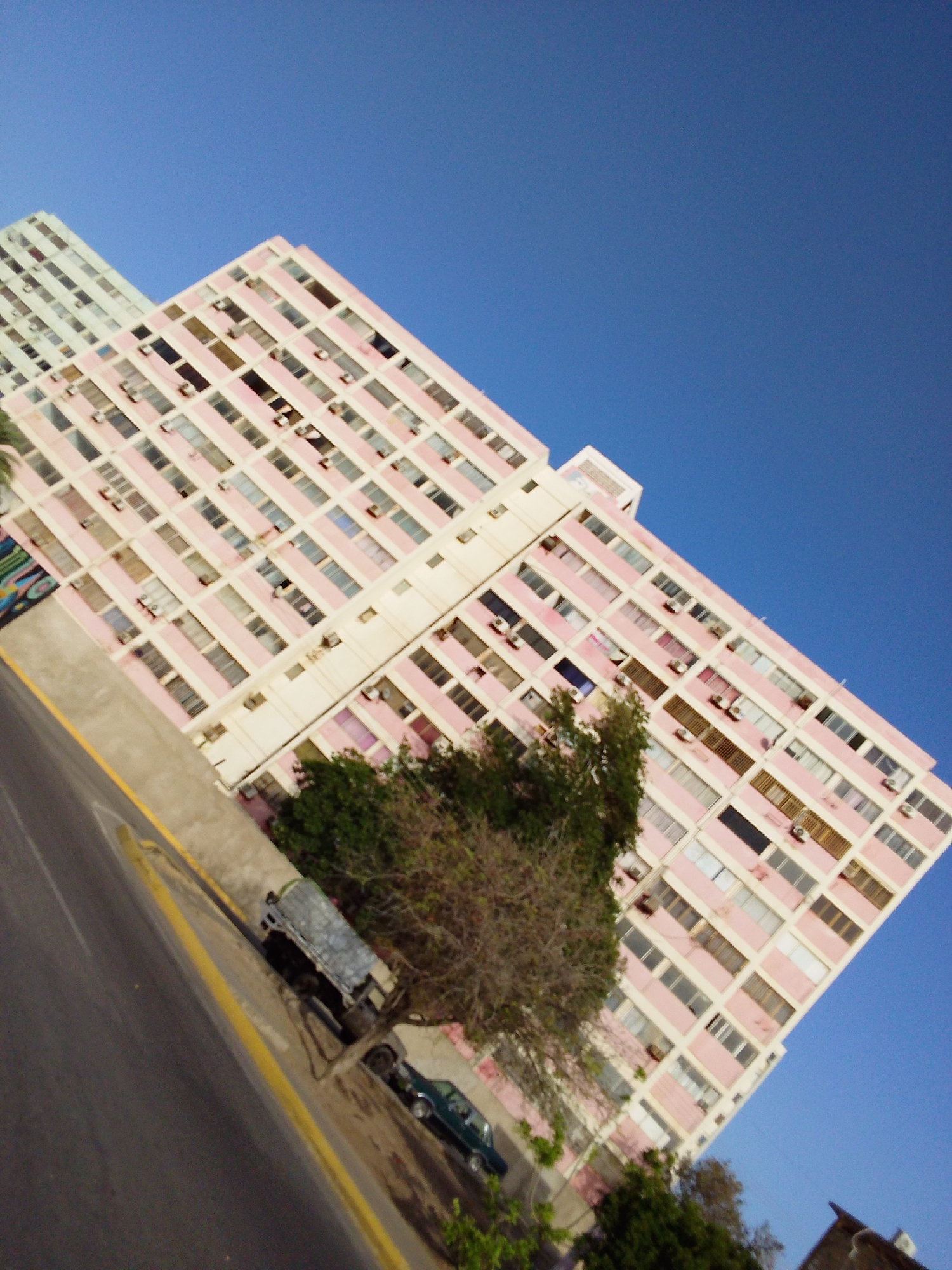 The image captures a skewed view of an urban landscape dominated by two tall residential buildings under a bright blue, cloudless sky. The perspective is tilted diagonally, causing the buildings and ground to appear slanted upwards from left to right.
The most prominent building is a multi-story apartment complex, predominantly pink and white, occupying the center and right side of the frame. It features numerous rectangular windows and balconies, many equipped with visible air conditioning units. The building's facade is characterized by a grid-like pattern of pink horizontal bands separating floors, interspersed with white vertical elements. To the left and slightly behind this building, another similar multi-story structure, primarily white or light grey, is partially visible.
At the bottom of the image, a paved road with a distinct yellow dividing line runs diagonally. On the side of the road, a dark-colored pickup truck with a covered, possibly canvas, bed is parked. Further down the street, a smaller dark-colored car is also parked, partially obscured by lush green trees and bushes that line the roadside and grow around the base of the buildings. The vegetation includes a mix of leafy trees and shrubbery.
The scene depicts a static moment with no overt human activity. The bright light and clear blue sky suggest it is daytime, likely mid-morning or afternoon. There is no visible text in the image.