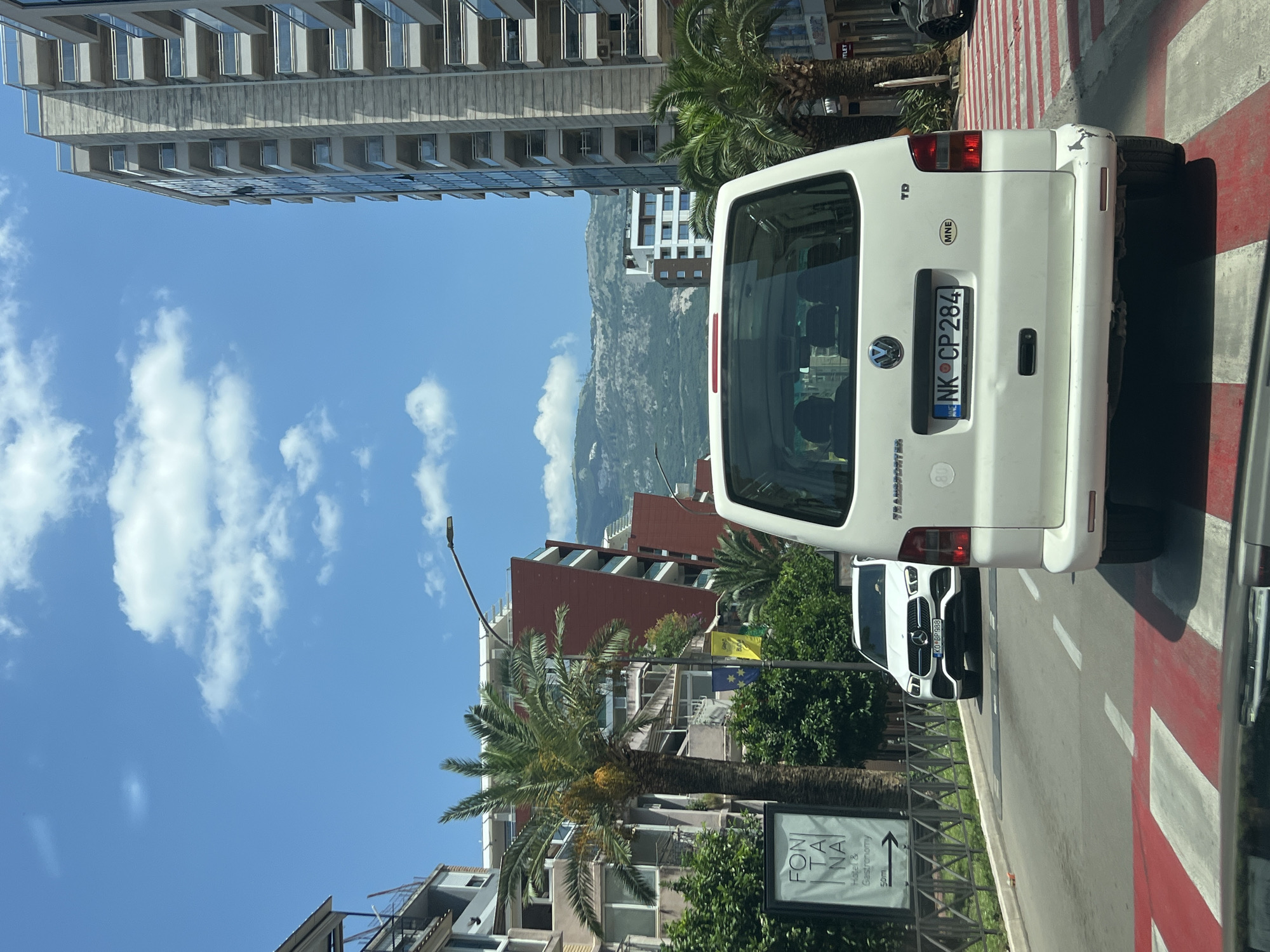 The image shows a rear view of a white Volkswagen Transporter van driving on a city street in Budva, Montenegro. The van has a license plate that reads "NK CP 284" and a "MNE" sticker indicating its origin. The van is in the right lane of a road with a red and white striped pedestrian crossing in the foreground. To the left of the van, a white Mercedes-Benz SUV is visible in the left lane. The street is lined with palm trees and modern buildings, including a tall, multi-story building on the right. In the background, mountains can be seen under a blue sky with scattered clouds. A sign for "FONTANA Hotel & Gastronomy" is visible on the left side of the street. The scene appears to be a sunny day, likely in a tourist area given the architecture and foliage.