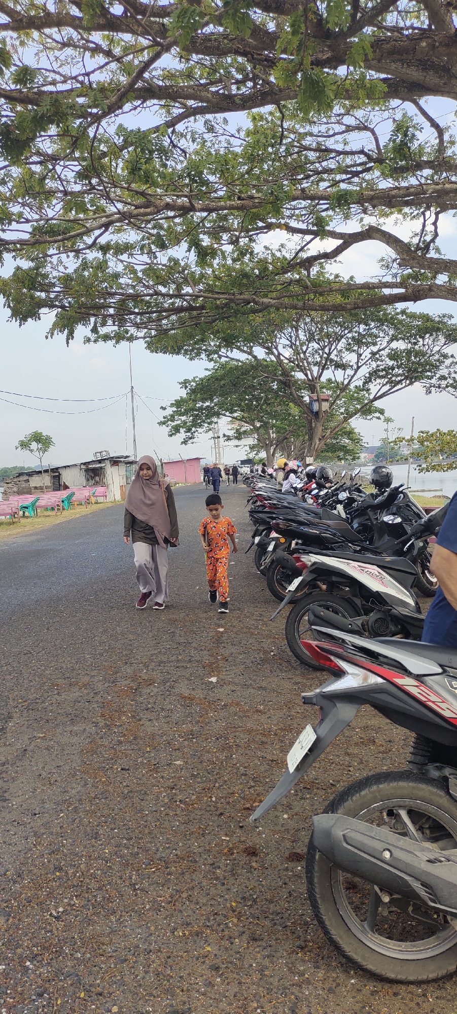 A woman in a brown hijab and loose-fitting pants walks on an asphalt road, accompanied by a young boy in an orange, patterned outfit. The boy is walking slightly ahead of the woman, carrying a tablet. They are walking towards the viewer, on a road lined with parked motorcycles. In the background, there are several buildings, some with corrugated metal roofs, and pink and green seating. Large trees with sprawling branches overhang the scene, and a body of water is visible in the distance on the right. The sky is overcast. The location appears to be a waterfront area, potentially a park or recreational space, in the city of Lhokseumawe, Indonesia, given the presence of parked motorcycles and the architectural style of the buildings.