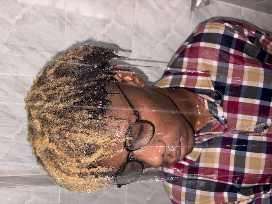 An individual, identified as male with dark skin, is depicted indoors under streams of falling water. The subject has short, curly hair, dyed blonde or light brown at the front, transitioning to dark brown or black at the back, heavily saturated with water droplets. Dark-rimmed eyeglasses are worn, obscured by water. The individual's head is tilted slightly down and to the left, with eyes appearing closed or cast downwards. The facial expression is neutral. A plaid button-up shirt in shades of red, white, blue, and tan/gold is worn, clinging to the torso due to wetness. Water is visibly running down the hair, face, glasses, and shirt. The background consists of light-colored, possibly gray or off-white, tiled walls, indicative of a bathroom or shower environment. The scene is located in Igboefon, Nigeria.