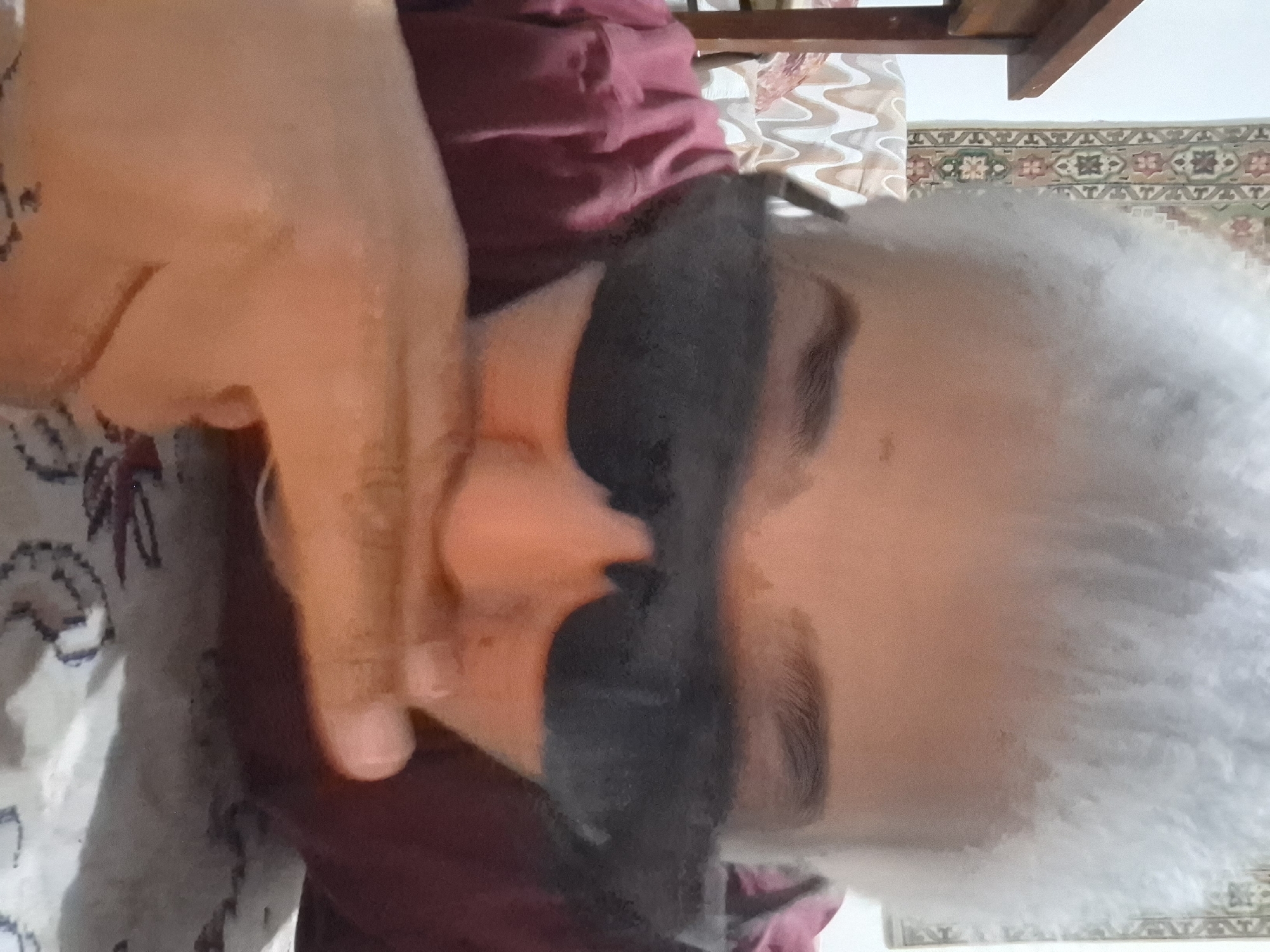 The image shows a close-up, waist-up shot of a person wearing dark sunglasses and a maroon or dark pink shirt. Their hand is raised to their face, with fingers touching their mouth. The person has light-colored, possibly gray or white, hair. The background consists of a patterned textile, possibly a rug or tapestry, hanging on a wall, and a portion of a wooden table on the left. A patterned sofa or chair is visible behind the person. The scene appears to be indoors, in a domestic setting. The location in Almaty, Kazakhstan, is not discernible from the image content.