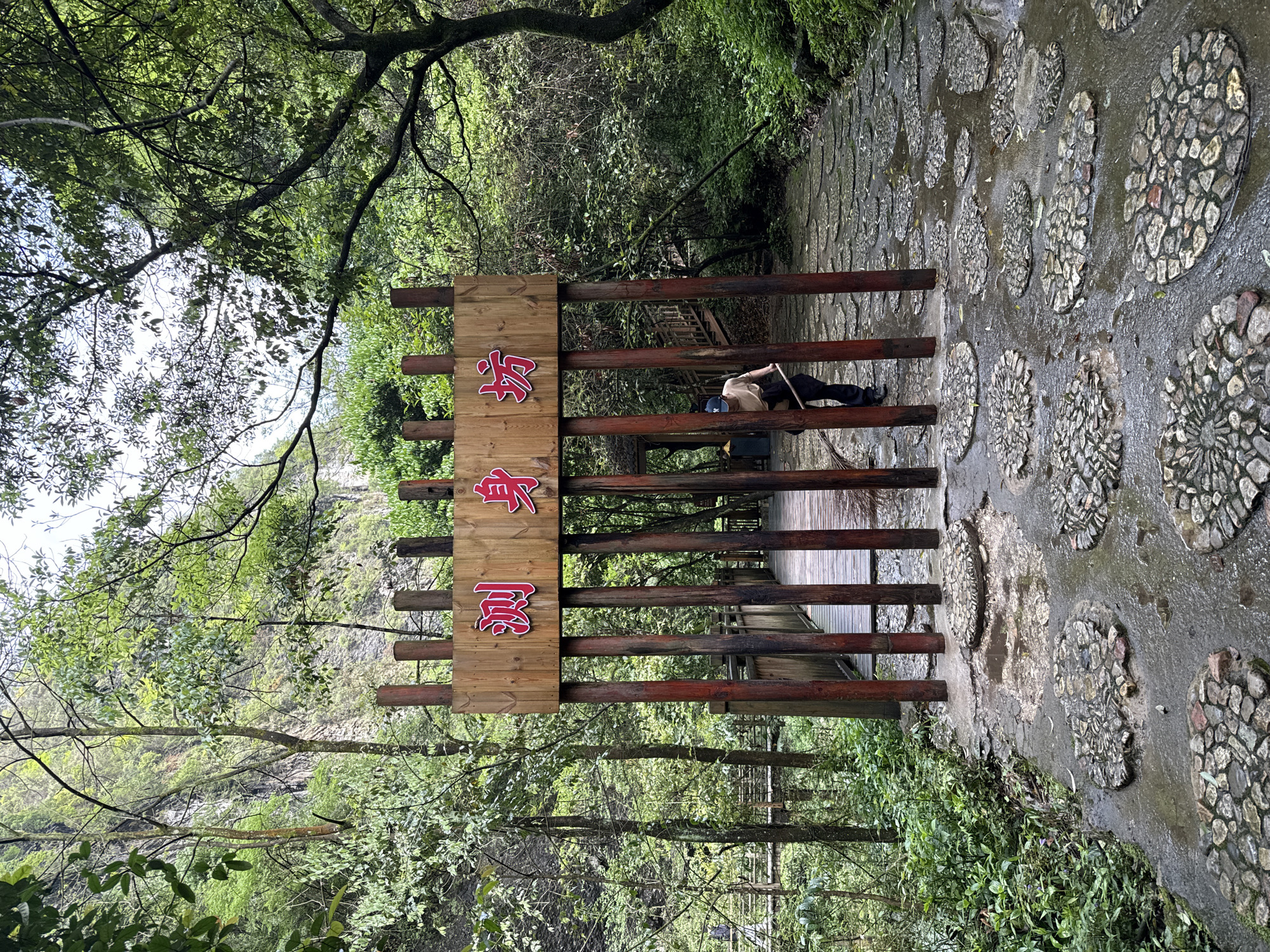 The image shows a scene in Zhangjiajie City, China, featuring a wooden gate or fence with Chinese characters in red. A person is lying across the fence, seemingly resting. The environment is lush with greenery, indicating a natural or park setting. A stone path with decorative rock patterns runs alongside the fence.