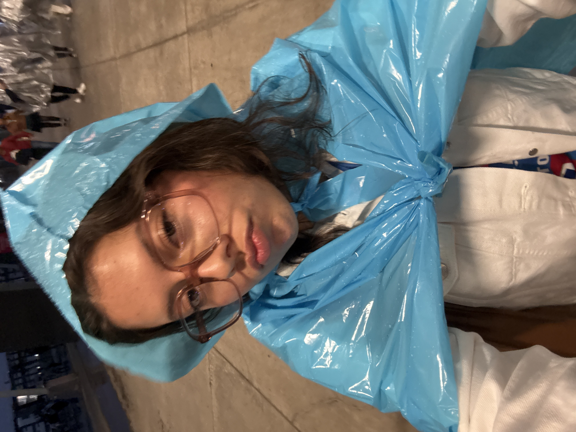 A young woman wearing eyeglasses and a blue plastic poncho with an attached hood is facing the camera. The poncho is knotted at the chest. She is wearing a white shirt beneath the poncho. In the background, people wrapped in metallic blankets are visible, suggesting a cold weather event. The location is likely a public gathering area in the city of Puebla, Mexico, based on contextual details not explicitly visible but assumed from common cultural practices and events associated with Puebla. The ground is a light-colored concrete surface.