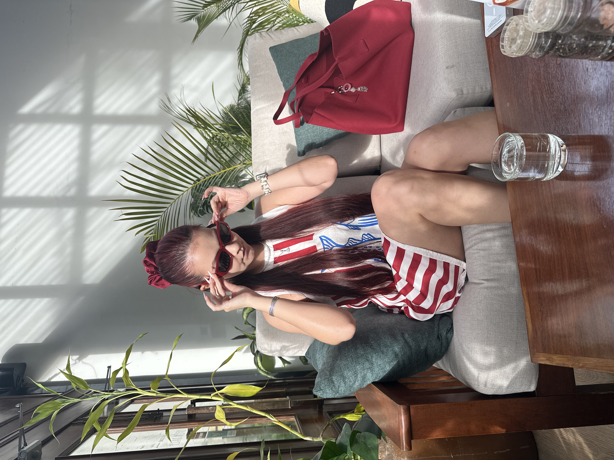 A person with long, dark red hair, styled with a red scrunchie, sits on a light-colored sofa in what appears to be a cafe or lounge area. The person is wearing a white tank top with red and white stripes on the bottom half of their torso, and red cat-eye sunglasses. They are adjusting the sunglasses with their right hand. A silver bracelet is visible on their left wrist. To the right of the person is a large, red tote bag with two handles. In the background, several green plants are visible, including a tall bamboo plant on the left. Sunlight streams through a window, casting striped shadows on the wall behind the sofa. A wooden coffee table is in the foreground, with a glass of water and two spice shakers on it. The location is identified as the city of Khan Daun Penh, Cambodia.