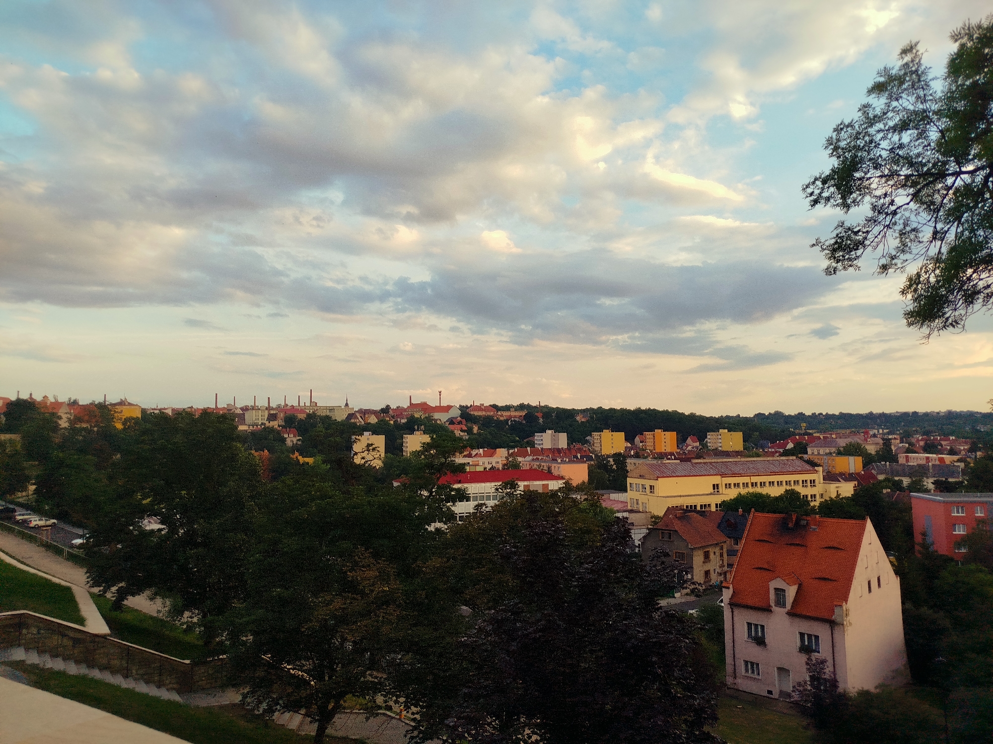 The image captures an elevated view of a cityscape with a dramatic sky. In the foreground, dark green trees partially obscure the scene, with a stone wall and steps leading down to a pathway on the left. Beyond the trees, a collection of buildings of varying sizes and colors makes up the urban landscape. Notably, a house with a prominent red tiled roof and dormer windows stands out. Further into the distance, a row of tall, slender industrial chimneys are visible against the horizon, suggesting a historical or industrial area. The sky is filled with large, fluffy clouds, some tinged with the warm hues of sunset, against a pale blue background. The overall impression is one of a tranquil evening over a European town.