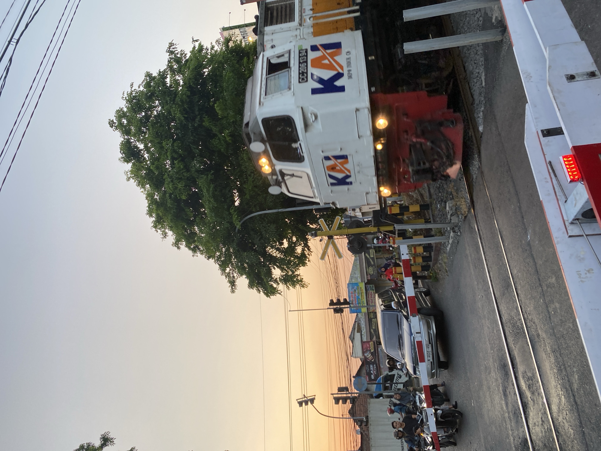The image depicts a train passing through a railway crossing in the city of Tebel, Indonesia. The sky is a gradient of pale blue to soft orange, indicating either dawn or dusk. On the right side of the frame, a white and orange train with the logo "KAI" is in motion, its headlights illuminated. The train's identification number, "CC 205 13 94," is visible on its side.
In the foreground, a paved road with visible railway tracks cuts across the image. A railway crossing barrier, painted red and white, is partially lowered, indicating that the crossing is active. On the left side of the crossing, several motorcycles and a dark-colored SUV are stopped, presumably waiting for the train to pass. A few people are visible on the motorcycles. A traffic light with multiple signals is mounted on a pole to the left of the crossing.
Further back, buildings with various advertisements are visible, suggesting an urban or semi-urban environment. A large tree with lush green foliage stands prominently behind the railway crossing barriers. Overhead power lines are strung across the upper portion of the image. The overall scene captures a moment of everyday life at a busy railway crossing during a transitional time of day.