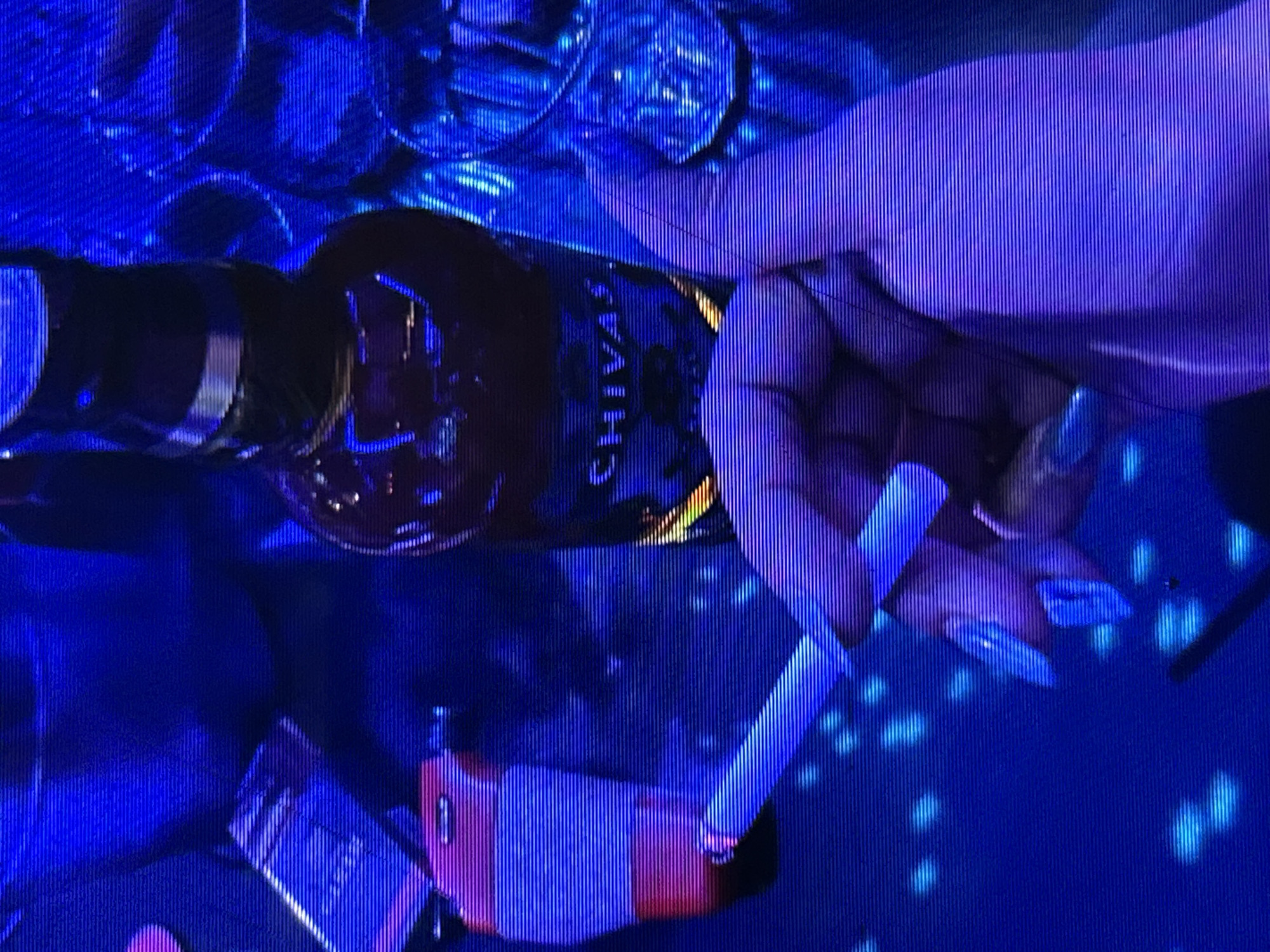 The image depicts a dimly lit scene, likely in an indoor setting such as a bar or club, given the presence of a bottle of Chivas Regal whiskey, glasses, and a cigarette. The dominant color is a deep blue, with streaks of purple and pink, suggesting the use of colored lighting. In the foreground, a hand with long, light blue acrylic nails holds a lit cigarette, the tip of which is glowing. To the left, a red and white cigarette lighter is visible. A pack of cigarettes rests on a surface, partially obscured. In the background, the Chivas Regal bottle is prominent, with its label clearly visible. Several glasses, some filled with liquid, are scattered around. The overall ambiance is one of nightlife and potentially a private gathering. The specific location in Depok, Indonesia is not identifiable from the visual elements.