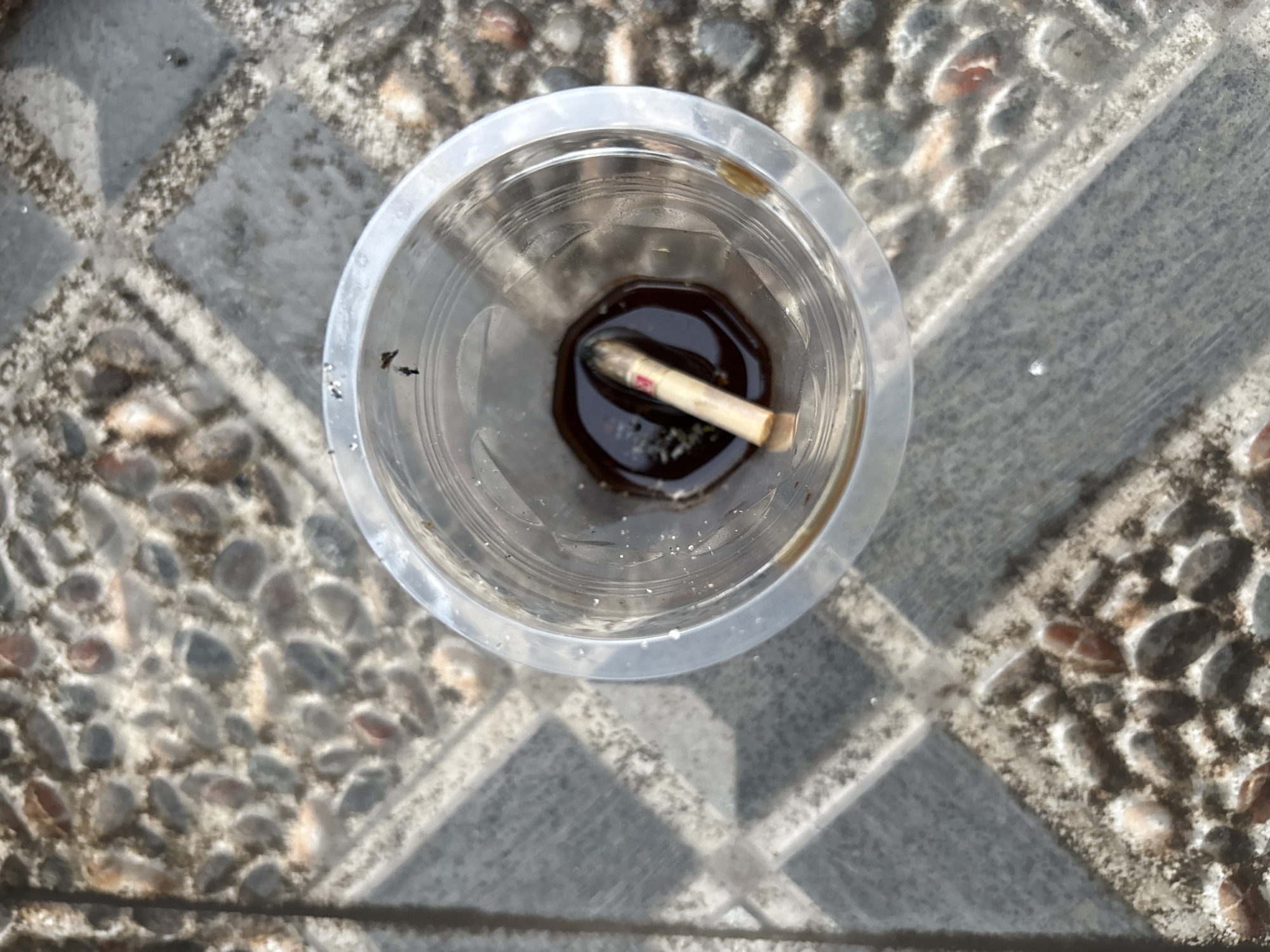 A close-up, overhead shot shows a used cigarette butt in a clear plastic cup filled with dark liquid, possibly water. The cup is placed on a textured outdoor surface made of interlocking grey and beige tiles with embedded pebbles. The scene appears to be in Palimanan, Indonesia. The lighting suggests it is daytime.
