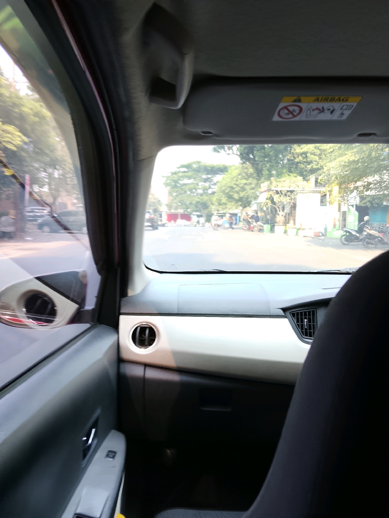 The image, taken from inside a vehicle, captures a street scene in RW 09 Kel. Oro Oro Dowo, Klojen Kota Malang, Indonesia. The interior of the car is visible, including a portion of the dashboard, a side door panel, and a seat. Outside the car, a moderately busy street is depicted with several motorcycles and a few cars. Buildings line the street, and trees are visible in the background. The lighting suggests it is daytime, possibly late morning or afternoon, with bright sunlight casting shadows. An "AIRBAG" warning label is clearly visible on the sun visor.