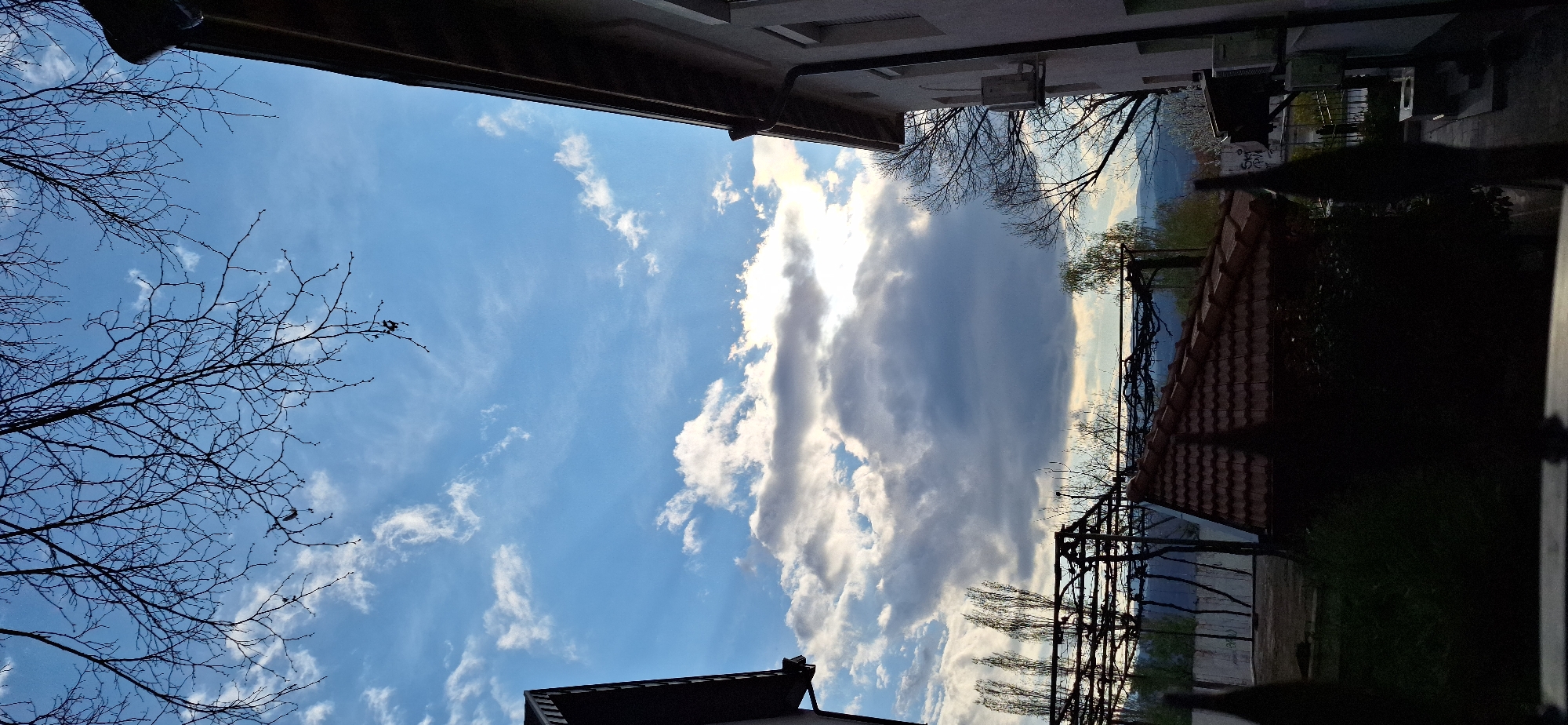The image shows a partly cloudy sky taken from what appears to be a balcony or window. Bare tree branches are visible in the lower left and right corners. In the background, a landscape with buildings and possibly mountains is visible. The sky is mostly blue with scattered white and gray clouds. The image is likely taken in an urban or suburban setting. The details are insufficient to specify Blagoevgrad, Bulgaria.