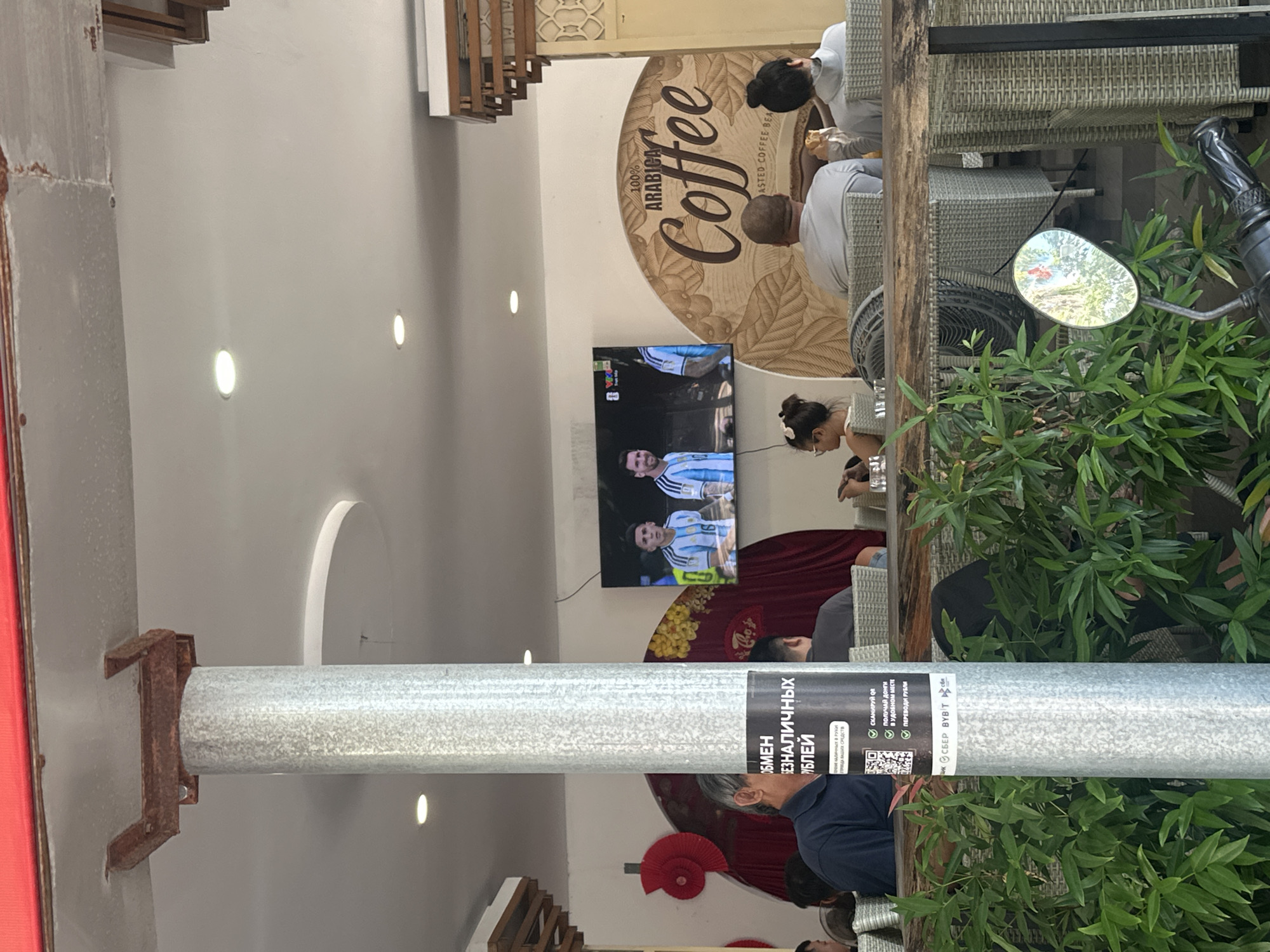 A wide shot captures an outdoor seating area of a cafe in Nha Trang Ward, Vietnam, with people dining and watching a football match on a television screen. A metal column stands in the foreground, partially obscuring a sign with Russian text and a QR code. Behind the column, several individuals are seated at tables, some eating and others engaged in conversation. A large TV screen displays a soccer game, with players in light blue and white jerseys visible. The cafe has a relaxed atmosphere, with rattan furniture and lush green plants decorating the space. A motorcycle mirror reflects the surroundings, including trees and buildings. The scene suggests a casual gathering, possibly during a sporting event.