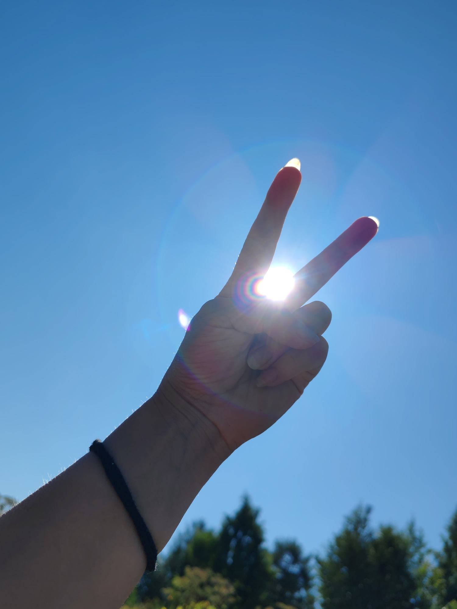 A hand is silhouetted against a bright blue sky, fingers extended in a V-shape, as if making a peace sign. The sun shines directly between the fingers, creating a lens flare and casting a bright glow. In the background, the tops of green trees are visible against the sky. The hand wears a dark bracelet on the wrist. The overall impression is a sunny day with clear skies. The location is not specified in the image or its context.