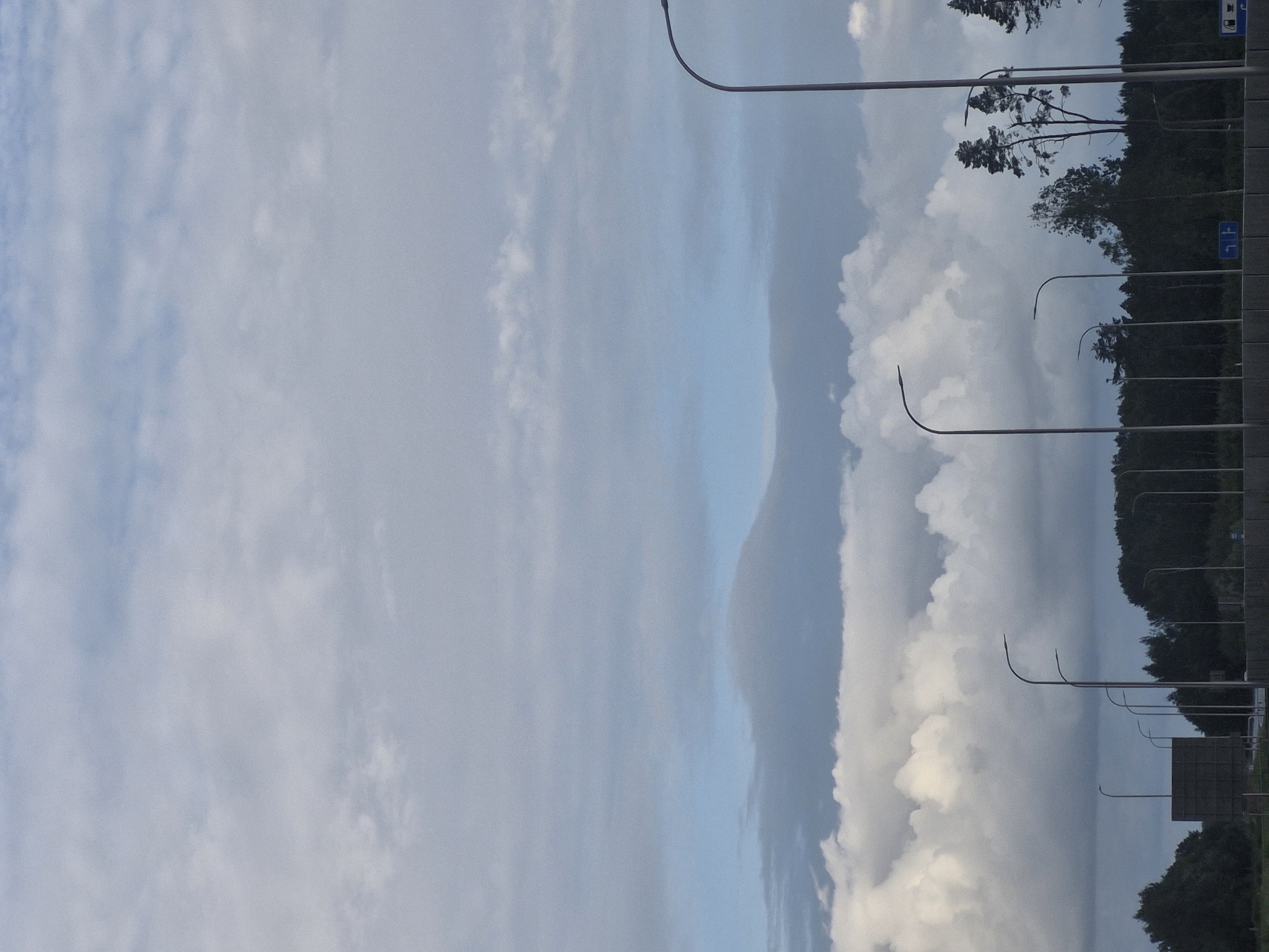 The image depicts a view from a road, likely a highway, with a line of streetlights and a dense forest in the background. The sky is filled with a dramatic cloudscape, featuring both wispy cirrus clouds at higher altitudes and large, puffy cumulus clouds closer to the horizon. A dark, layered cloud formation is visible in the mid-ground, adding depth to the sky. The trees in the background are dark green and form an unbroken line, suggesting a natural or park area. Several streetlights, with their distinctive curved arms, are spaced along the road. A large, dark, rectangular structure, possibly a billboard or sound barrier, is visible on the left side of the frame. The overall scene is overcast, with muted colors, and the lighting suggests it is either early morning or late afternoon. The location is identified as being in the vicinity of Jenči, Latvia.