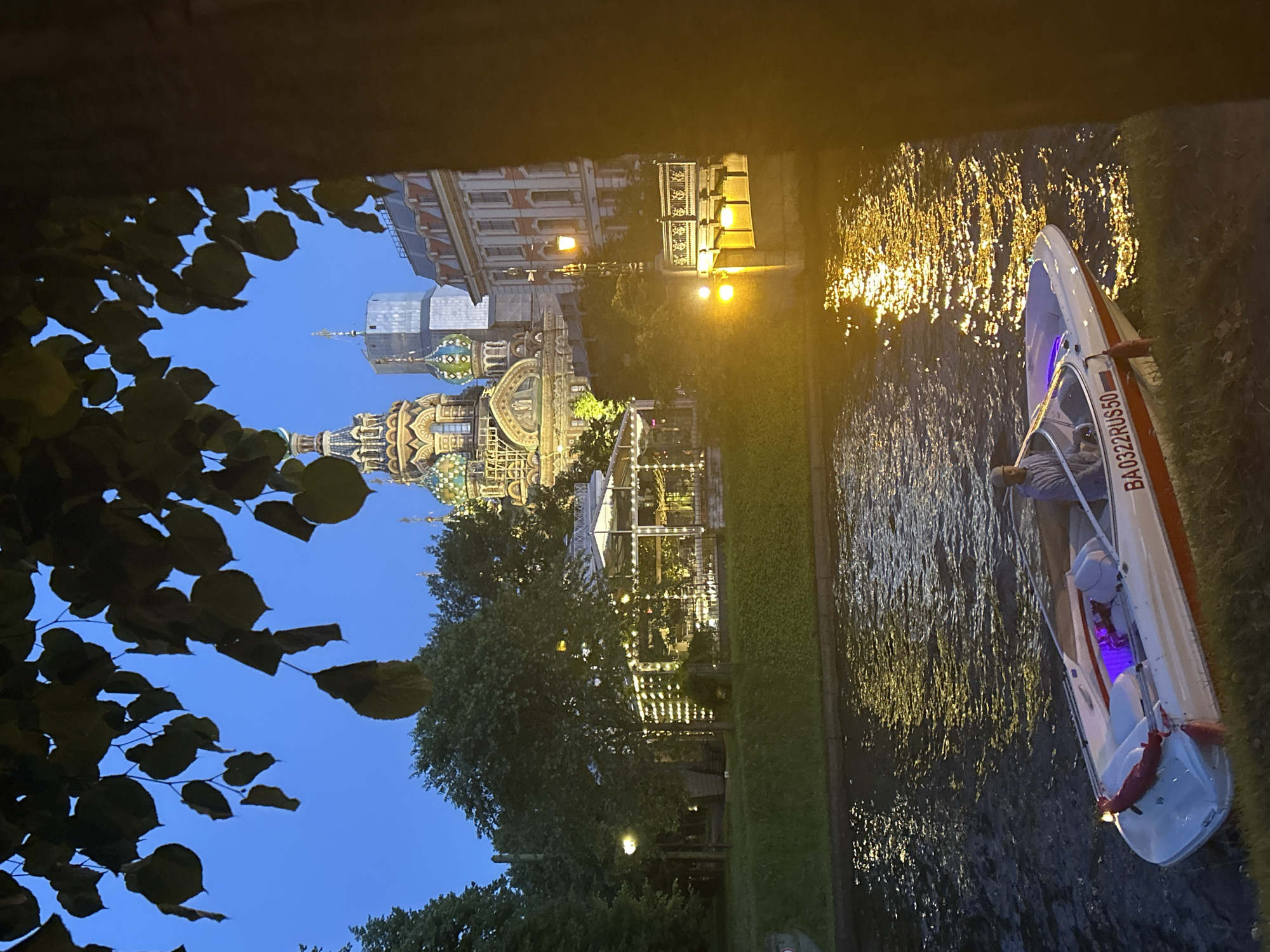In an urban canal scene at dusk, a white speedboat with red trim, bearing the registration "BA0322RUS50," glides near the bank. A single individual, wearing a grey sweater and a cap, is seated at the helm. The water's surface reflects the warm glow of streetlights and illuminated buildings, creating a shimmering effect. Behind the canal, a verdant grassy embankment is adorned with trees. Further in the background, a prominent ornate building with colorful domes and spires, likely a church, stands illuminated. Adjacent to it, a modern tall building and a red-roofed structure contribute to the city's architectural tapestry. Foliage from overhanging trees frames the upper portion of the image, obscuring parts of the sky and distant structures. The overall atmosphere suggests a tranquil evening in a city with a blend of historic and contemporary architecture.
